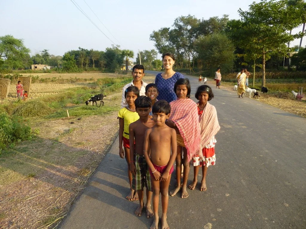 A group of children and two adults standing on a rural road, with trees, farm animals, and villagers in the background.