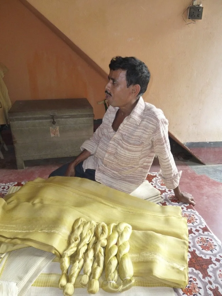 A man sitting on the floor next to a table with yellow silk fabric and silk threads, in a room with orange walls and an electrical outlet.