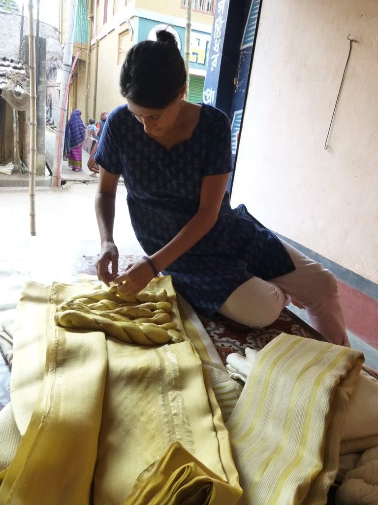 A woman kneels on a patterned rug, working with yellow fabric and twisted strands of yellow fabric, possibly preparing textiles, with a street scene visible in the background.