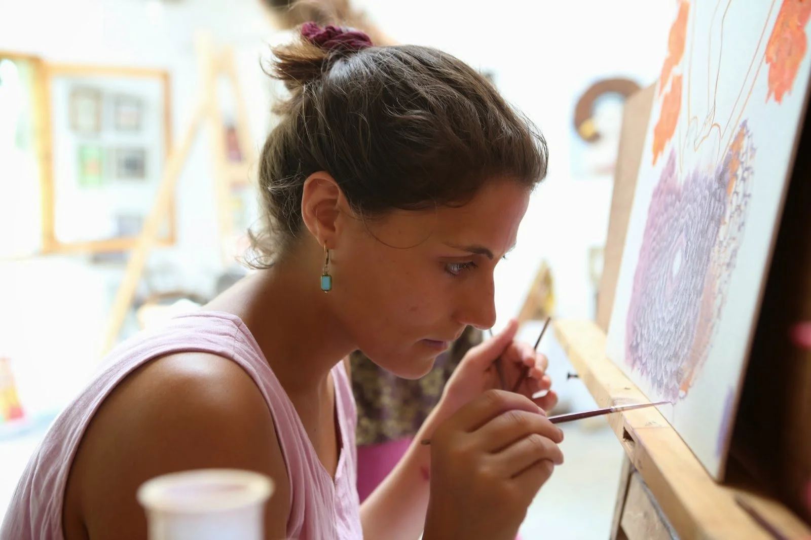A woman with brown hair tied up, wearing a pink sleeveless top and turquoise earrings, is carefully working on a painting on an easel, using a thin brush.