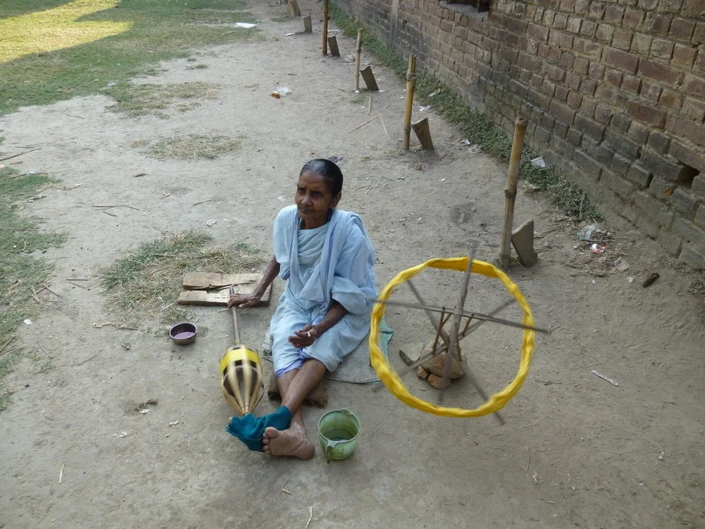 An elderly woman dressed in white sitting on the ground outdoors, surrounded by makeshift toys including a yellow ring-and-stick toy and a small spinning top, with two cups nearby, against a brick wall and a dirt pathway.