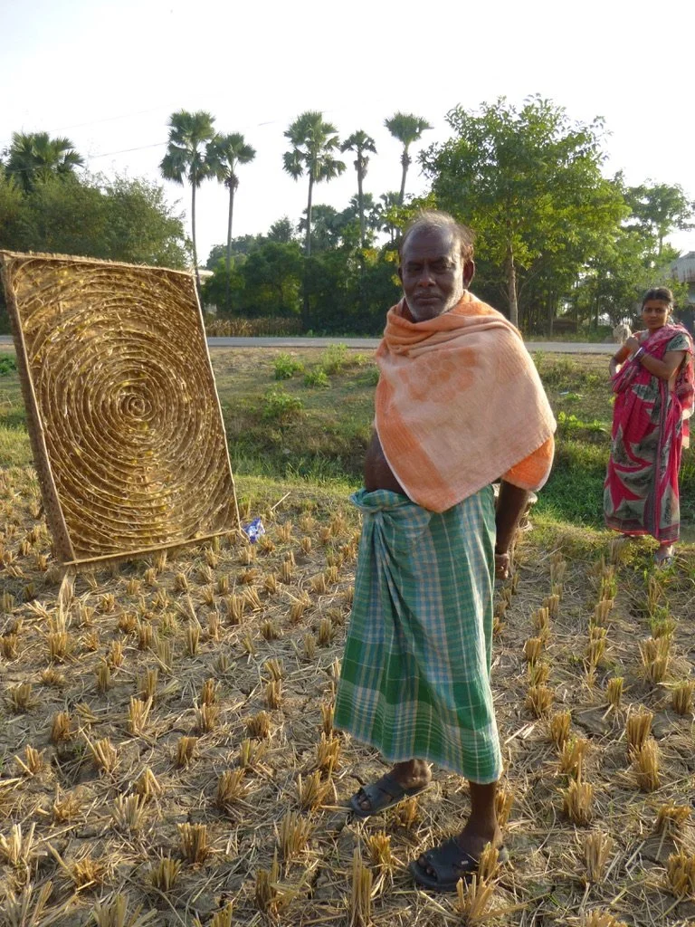 An elderly man in traditional clothing stands in a harvested rice field next to a large circular rice straw drying rack. A woman stands in the background among trees and palm trees, with a road visible behind them.