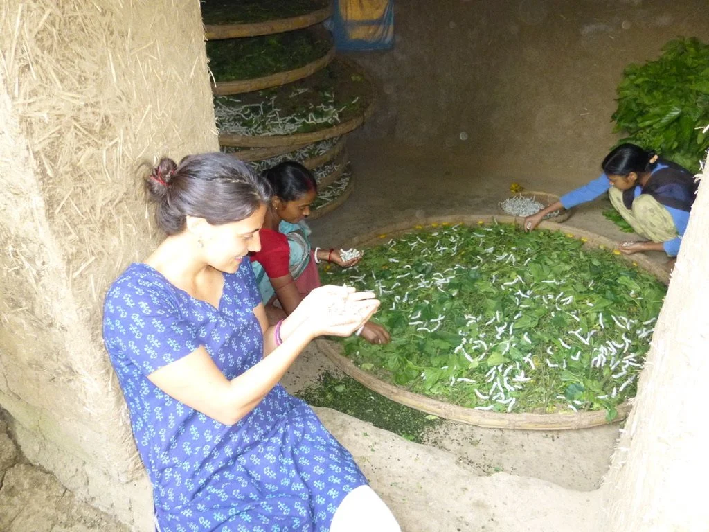 Four women sitting and squatting around large round trays filled with green leaves and white caterpillars, in a bamboo-walled room.
