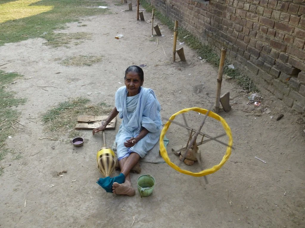An elderly woman sitting on the ground outdoors, with a spinning wheel and a mug nearby, next to a brick wall.