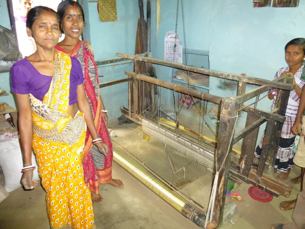 Four women standing inside a room with a large wooden weaving loom, some of them smiling at the camera.
