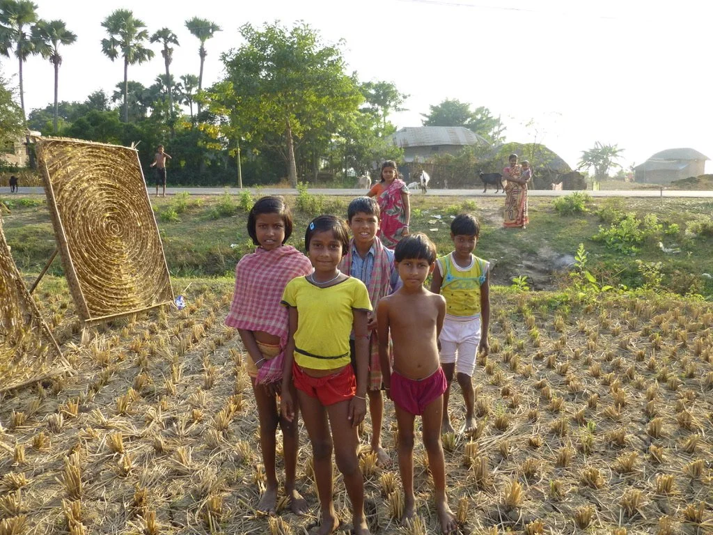 Group of children standing in a field with harvested crops in a rural area, with trees, houses, and some adults in the background.