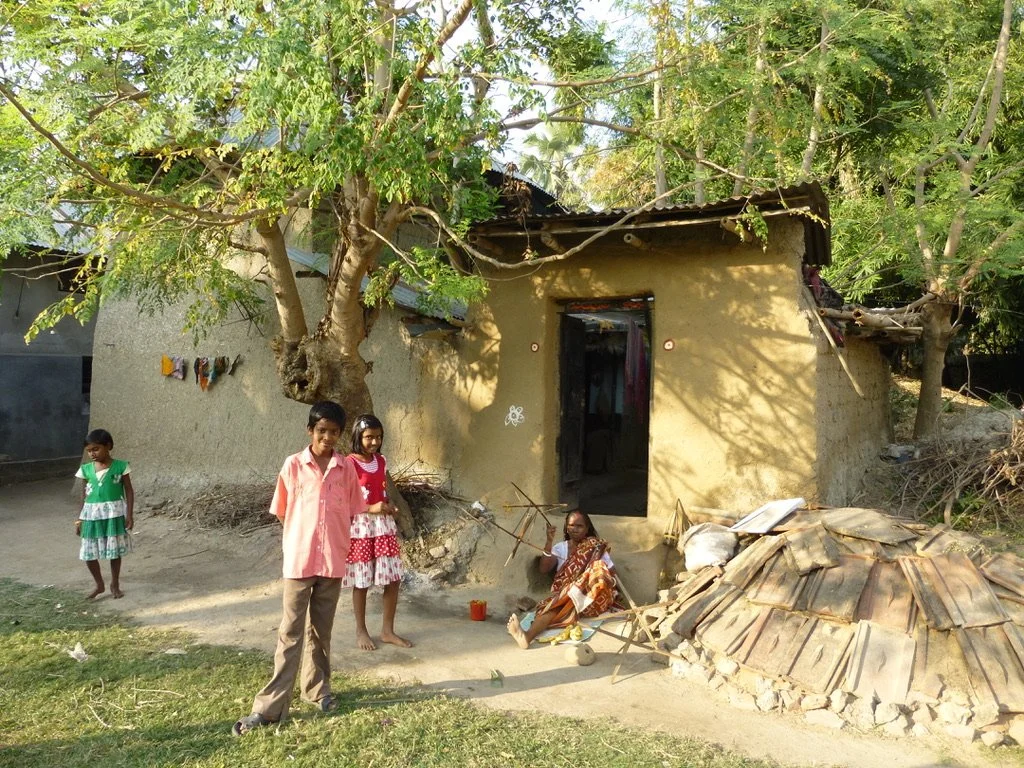 A rural scene with a small adobe house, a large tree in front, and four children standing outside. One woman is sitting near the house, and there is a pile of old roofing sheets nearby.