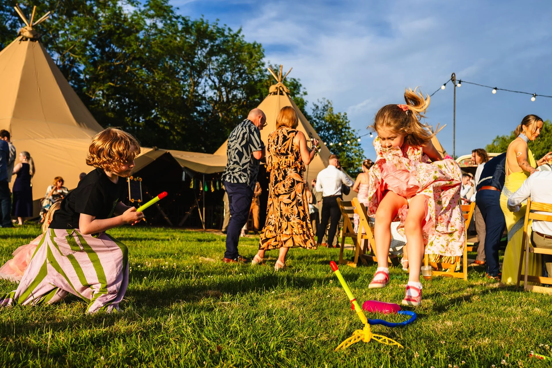 Children playing outdoors at a festival wedding in Cheshire, with teepees and string lights in the background, under a partly cloudy sky.