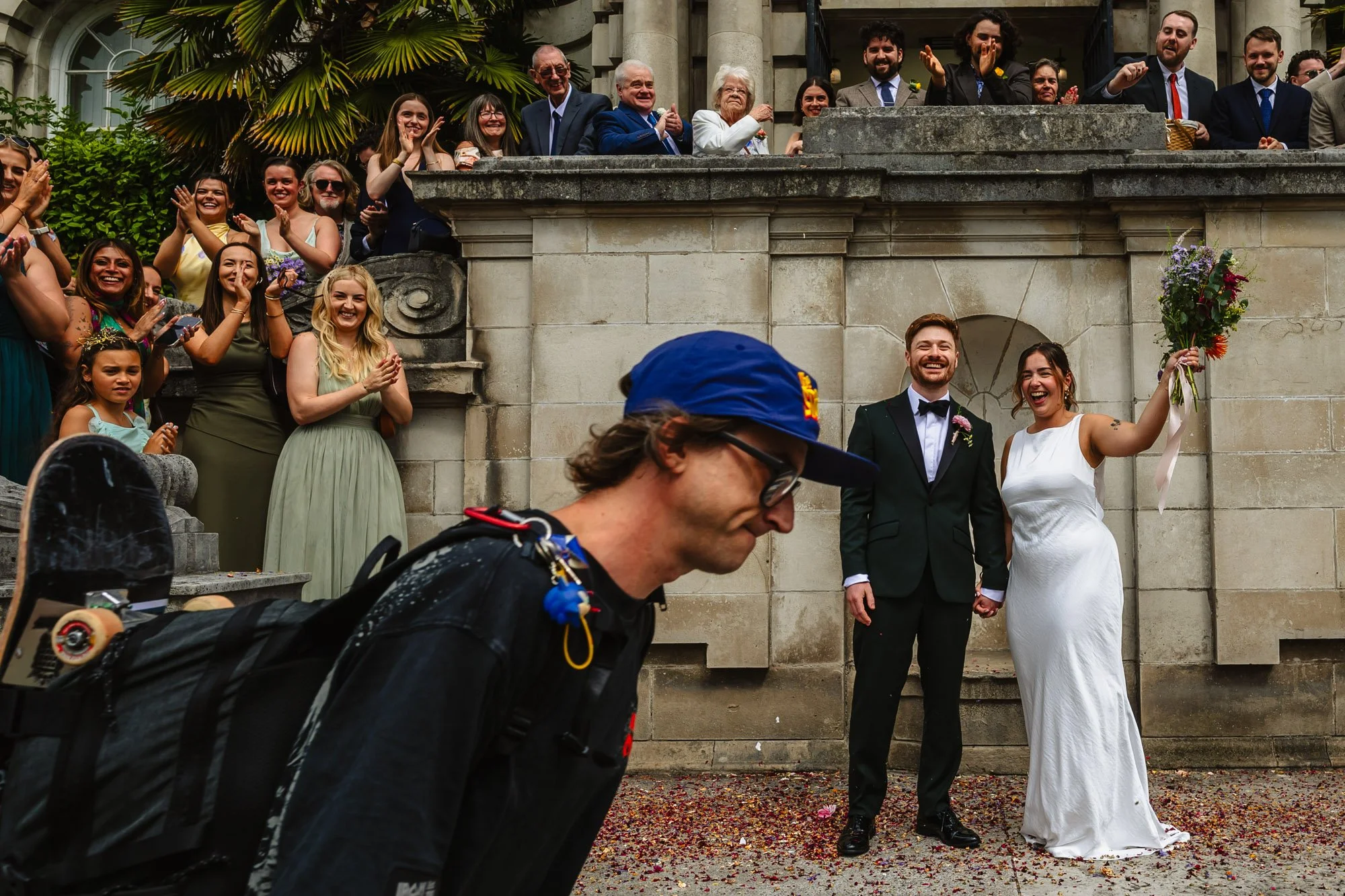 A reportage style photograph of a man in a blue baseball cap with a skateboard walking past a wedding party outside stockport town hall moments after a confetti throw
