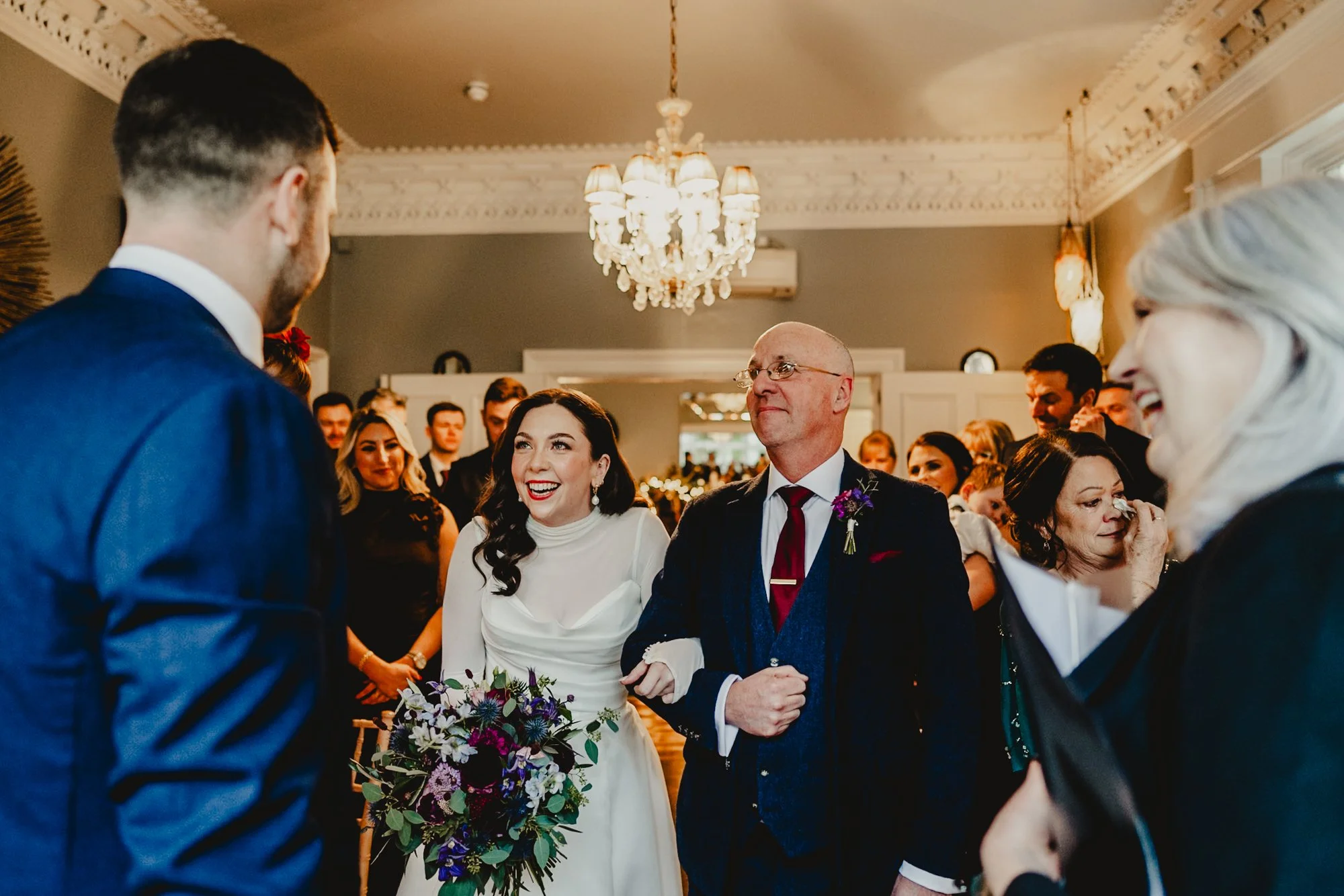 A wedding ceremony with a bride in a white dress holding a bouquet, walking arm-in-arm with her father. Guests are smiling and watching in a decorated indoor venue with a chandelier.