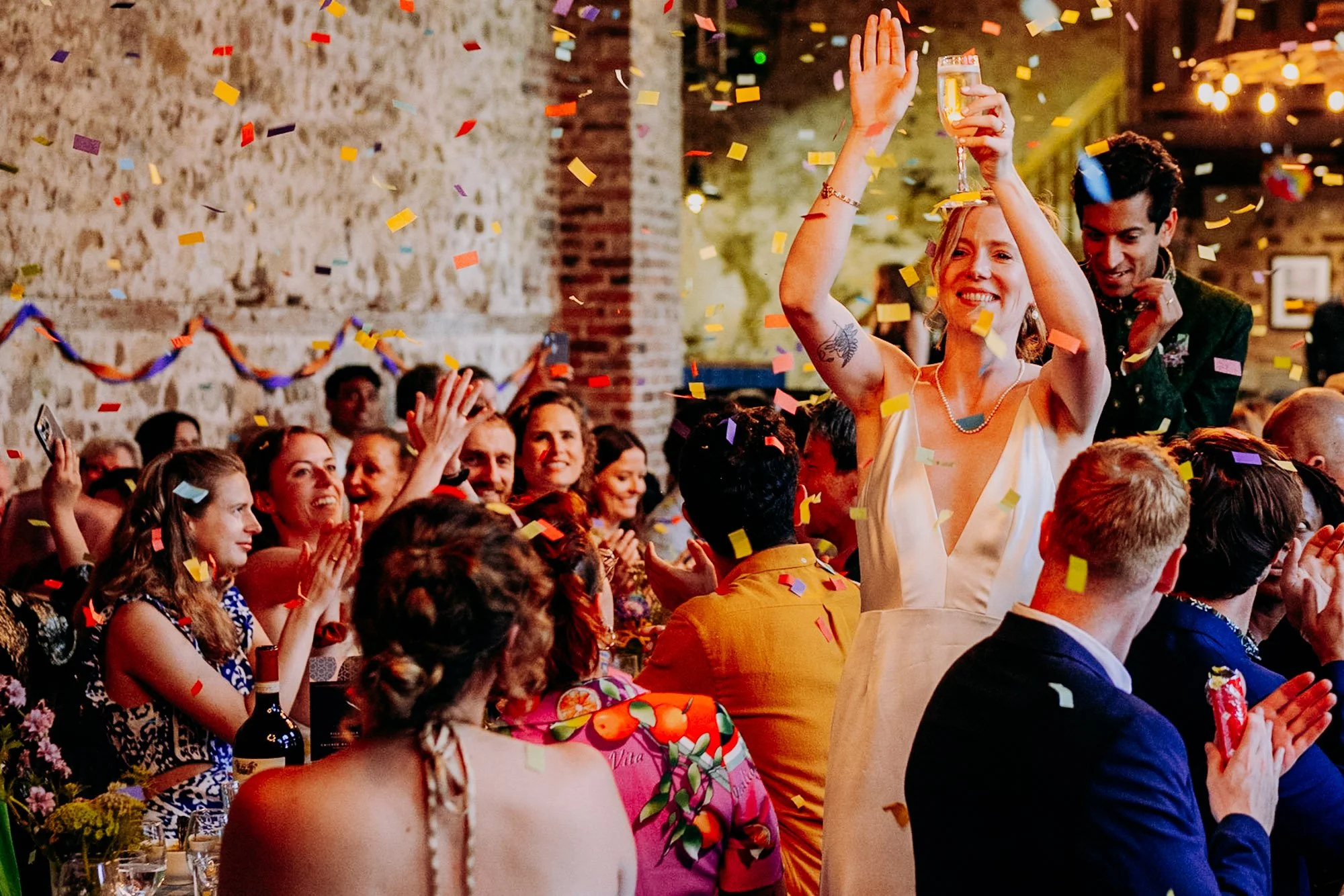 Photograph of a couple walking through long tables of guests at their wedding reception in a barn as confetti falls through the air. All the guests are applauding with their hands in the air.