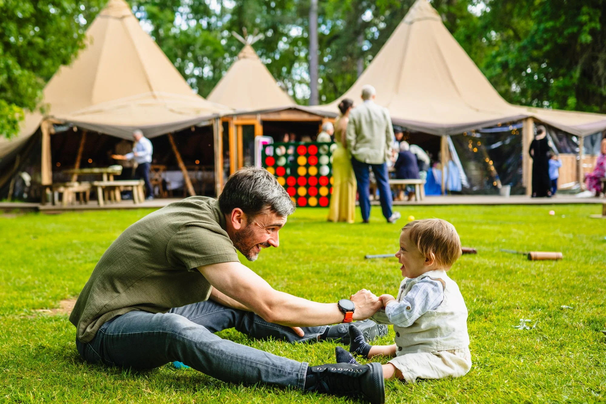 A man and a baby sit facing each other in front of the tipis at Whatton House during a wedding reception by documentary wedding photographer Caroline Goosey