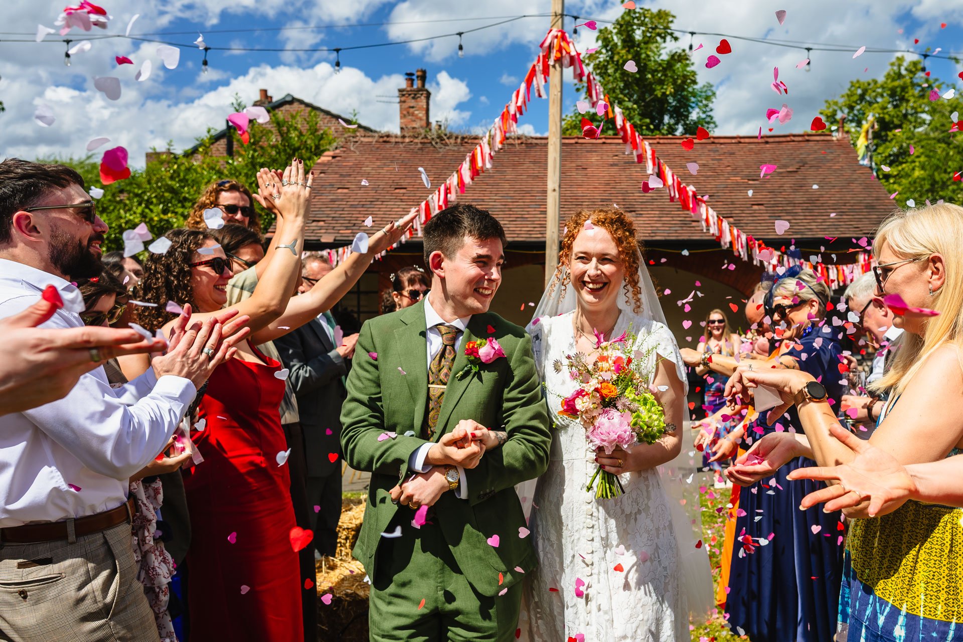 A bride and groom smiling as they hold hands, surrounded by guests at an outdoor wedding celebration with colorful confetti falling.
