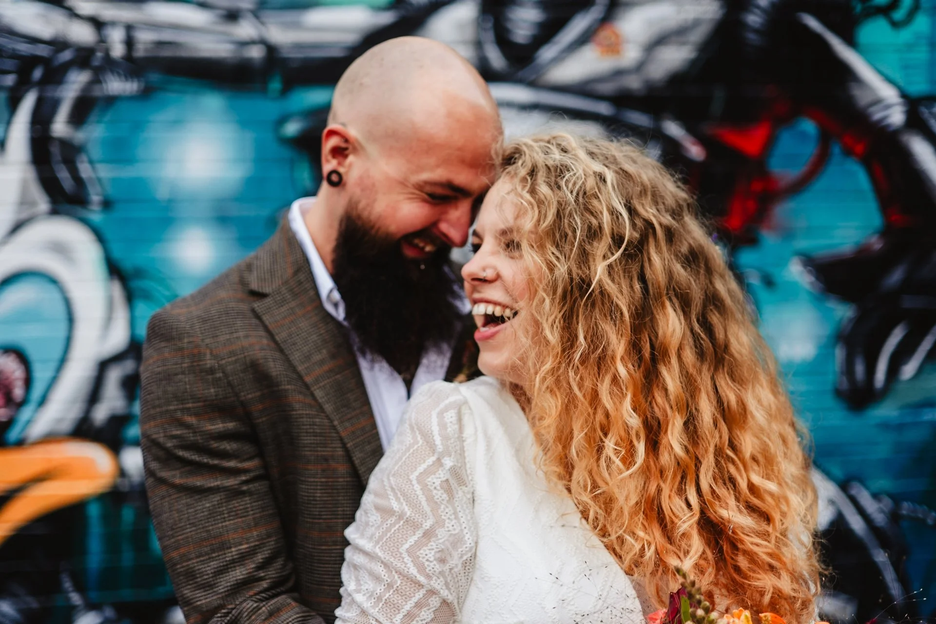 A couple smiling and leaning their foreheads together in front of a graffiti wall.