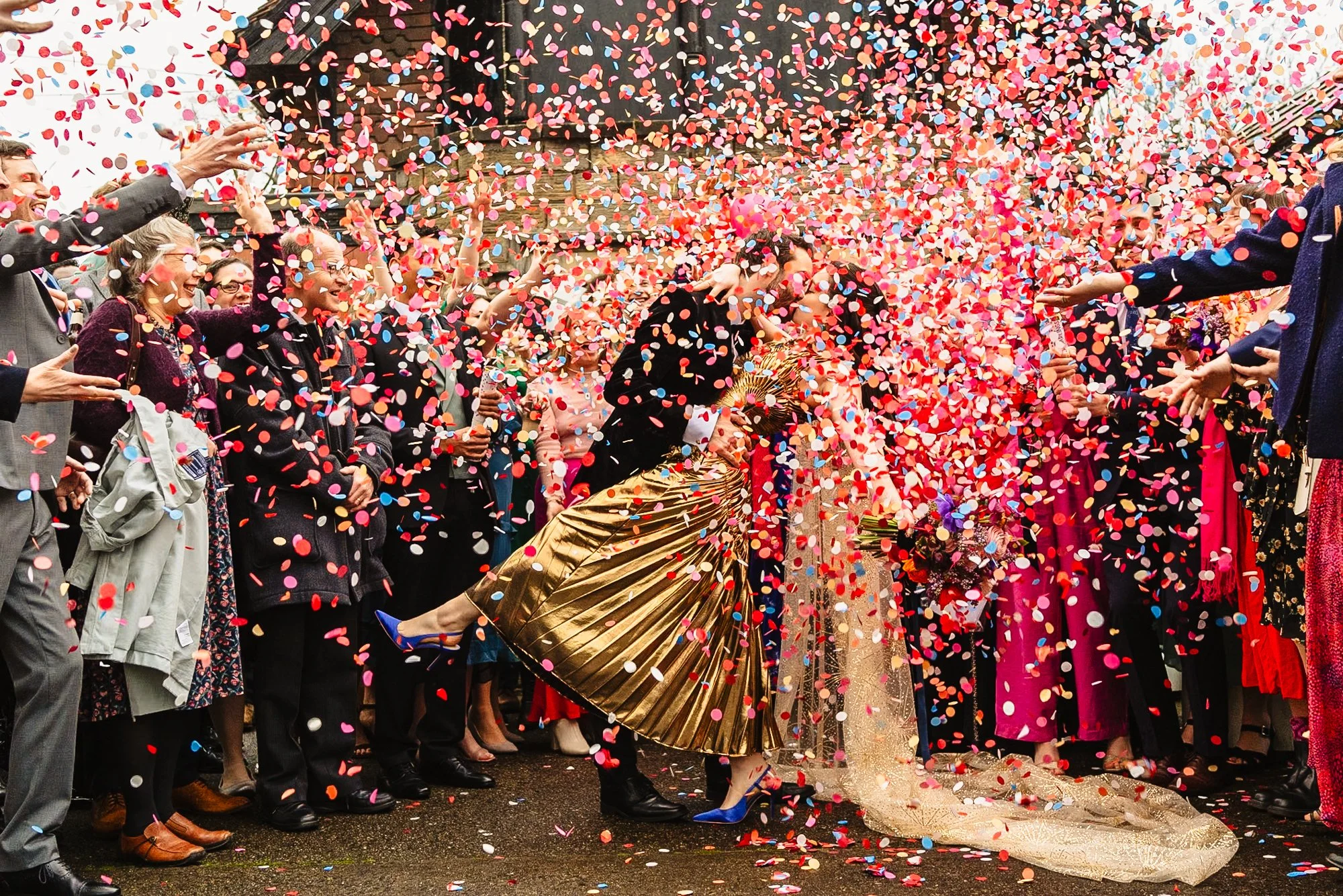 photograph of a huge cloud of pink and blue confetti being thrown over a couple kissing with their guests surrounding them