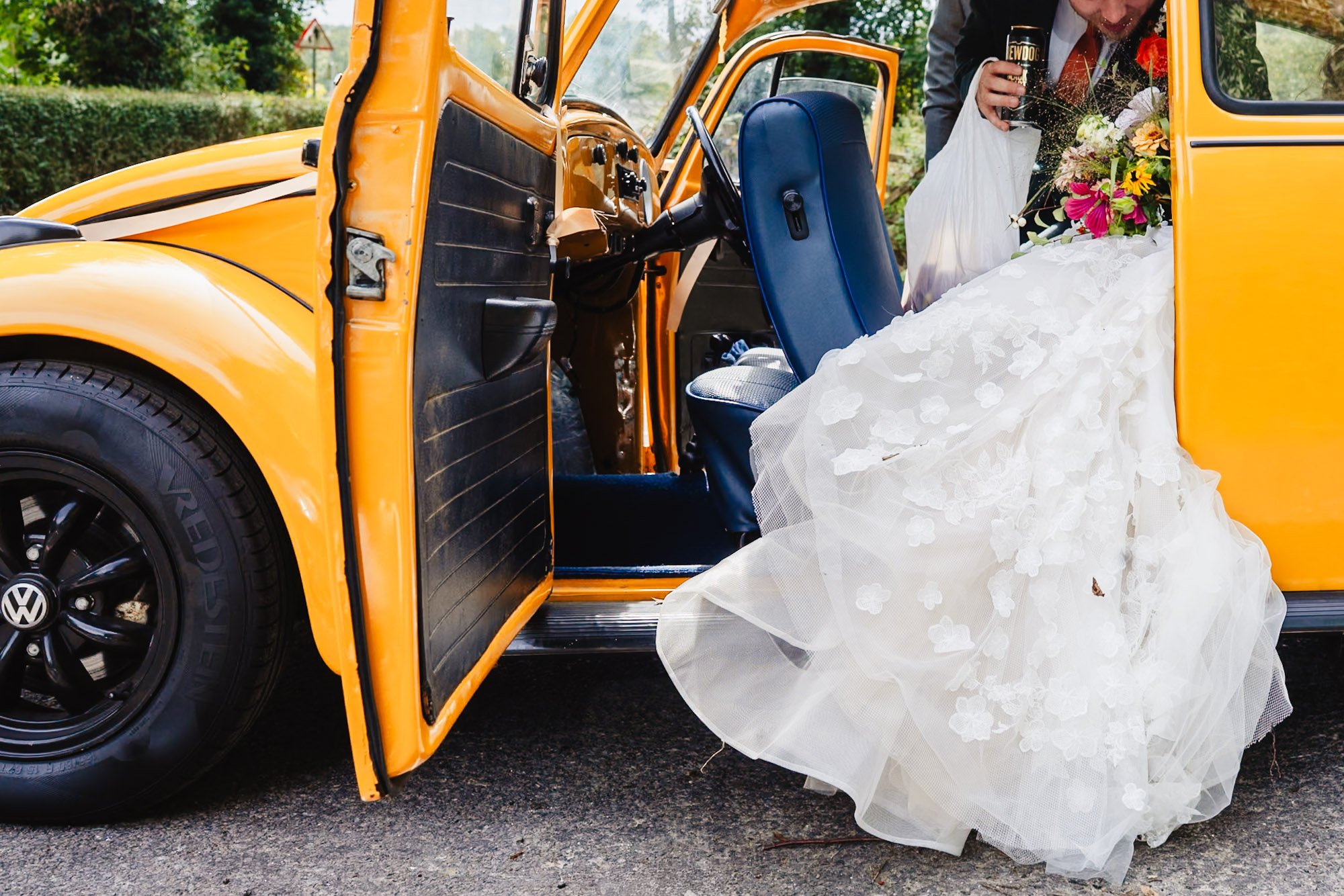 close up detailed photograph of a bride and groom getting in their vintage VW beetle at their wedding in Stockport. She is holding flowers, while he holds a carrier bag and a can of lager.