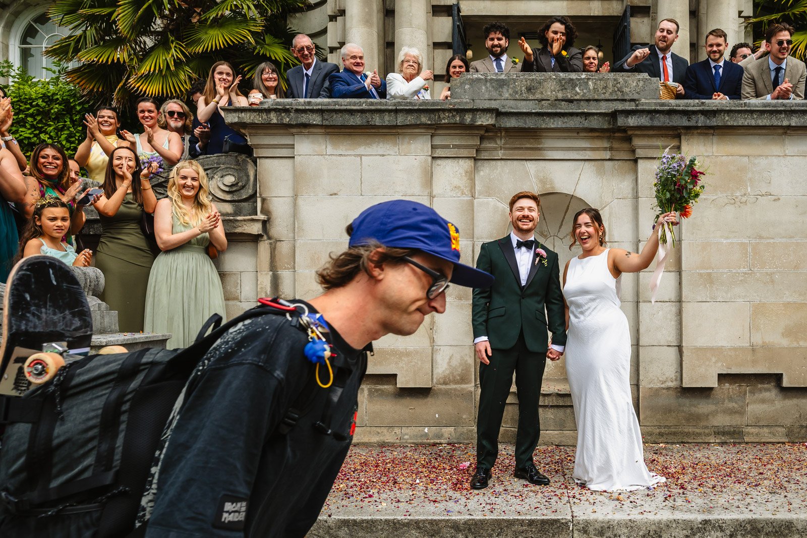 Photograph of a man in a blue cap, carrying a skateboard, walking through a confetti throw photograph outside Stockport Town Hall