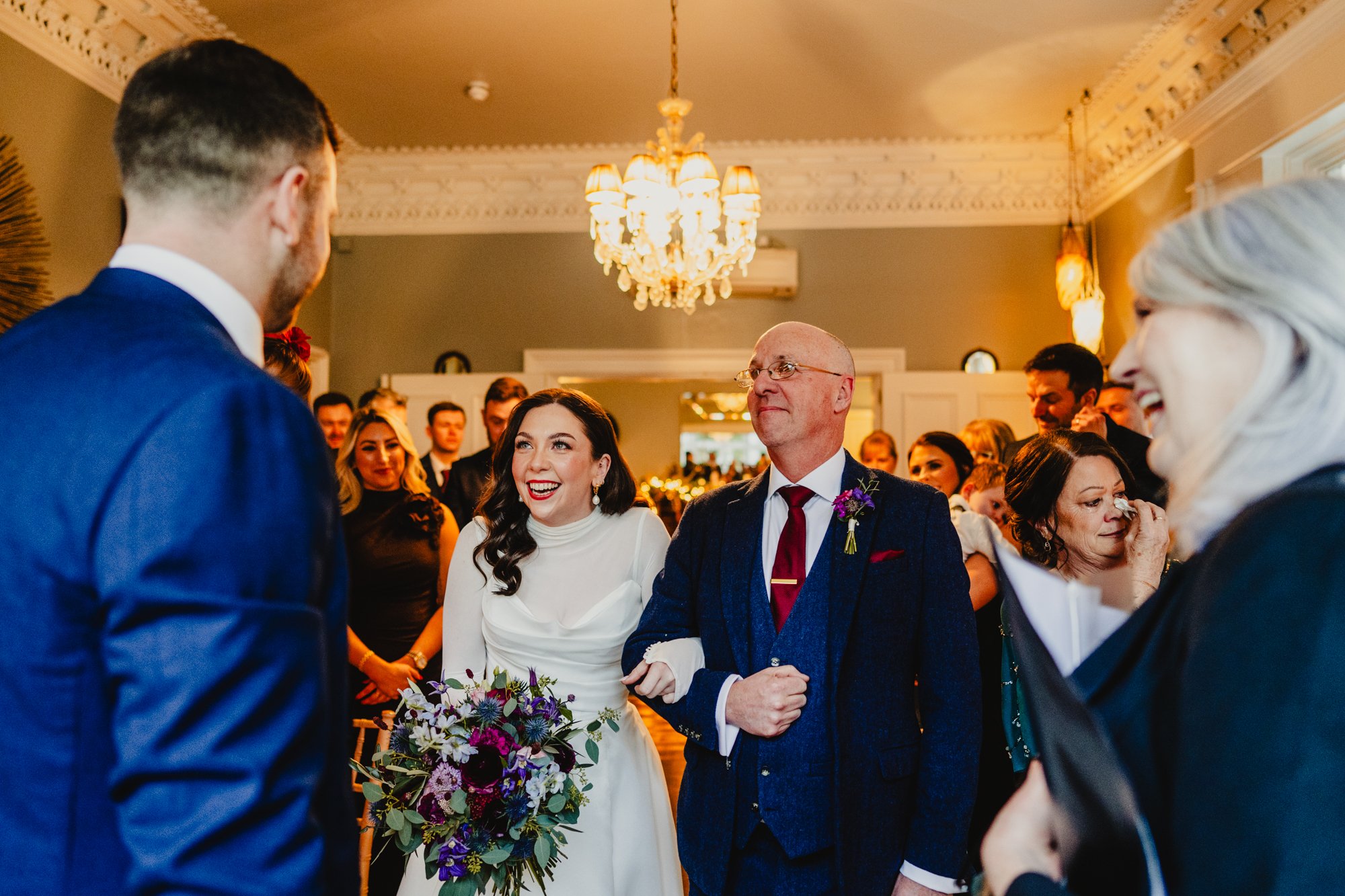 A bride in a long sleeved white gown is greeting her groom with a big smile on her face, and standing by her dad. All guests look a mx of happy and emotional. Photo taken at Didsbury House Hotel wedding venue near Manchester.