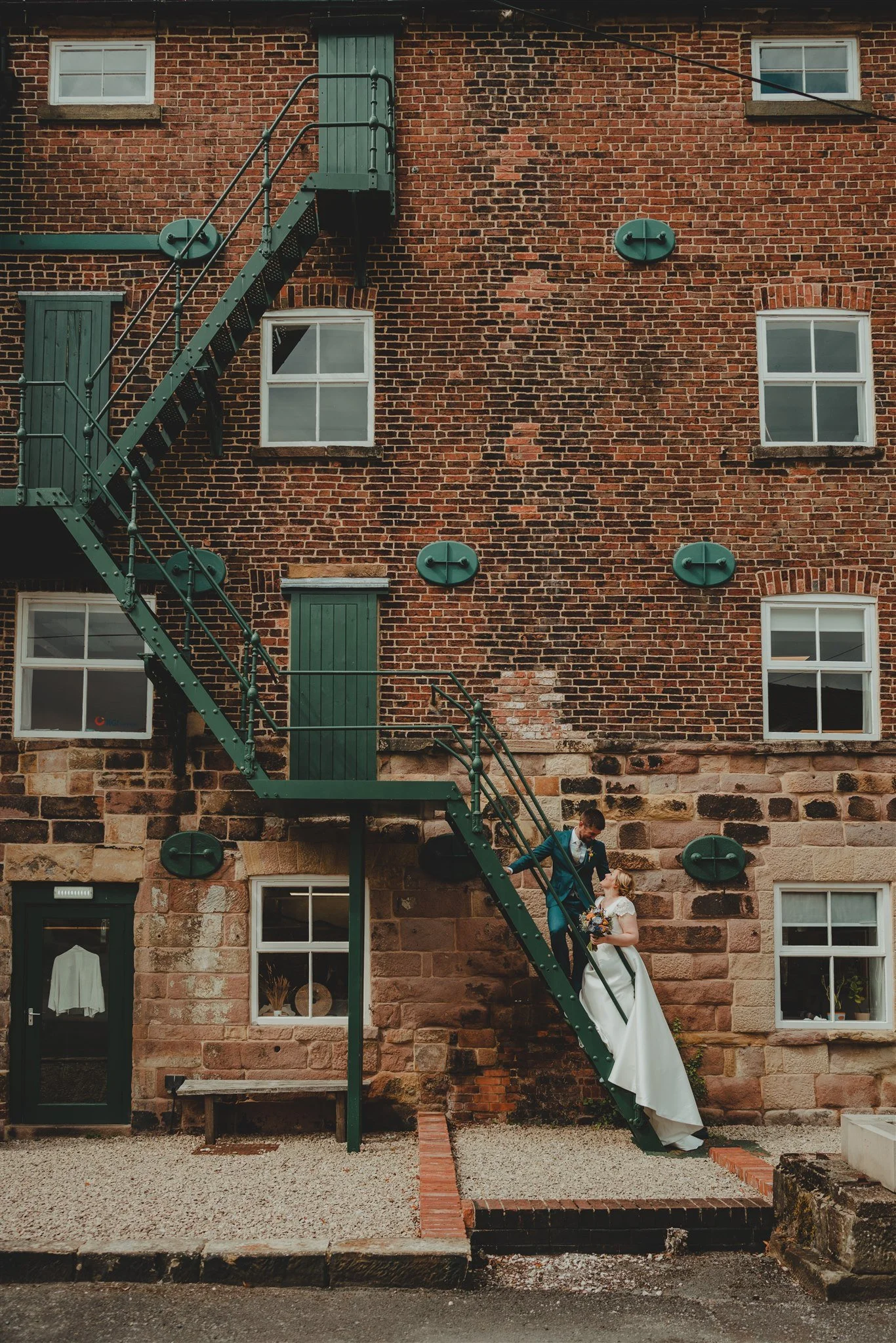 A couple dressed in wedding attire sharing a kiss on a spiral staircase outside a brick building with multiple windows. The wedding venue is Haarlem Mill, in Wirksworth, Derbyshire