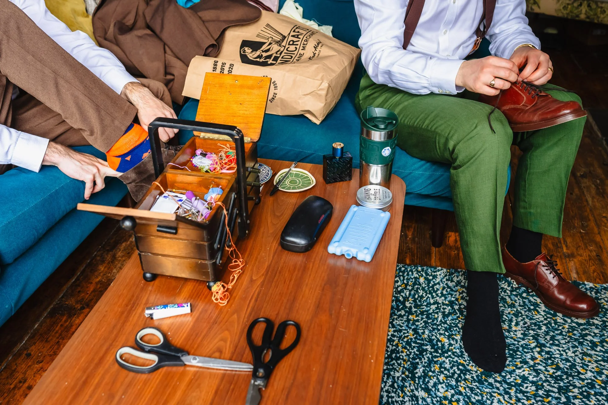  Two men wearing suits put their shoes on. They are sat at a table with a mid-century sewing box and a beer on it 