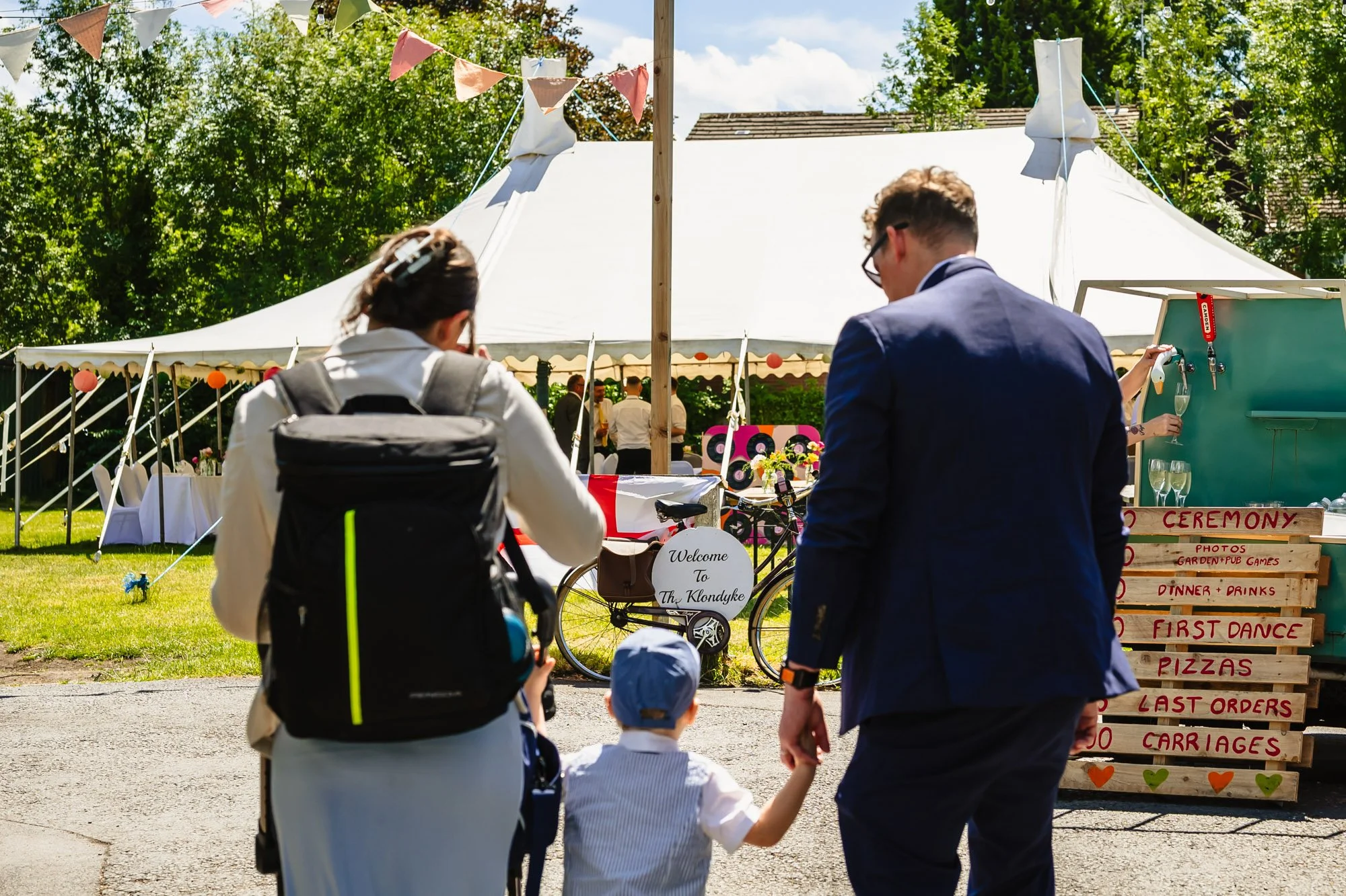  Photograph of two adults with a small child, entering the Klondyke in Levenshulme, Manchester, for a wedding. Visible in the image are the large marquee, mobile bar and a pallet painted with the order of the day. 