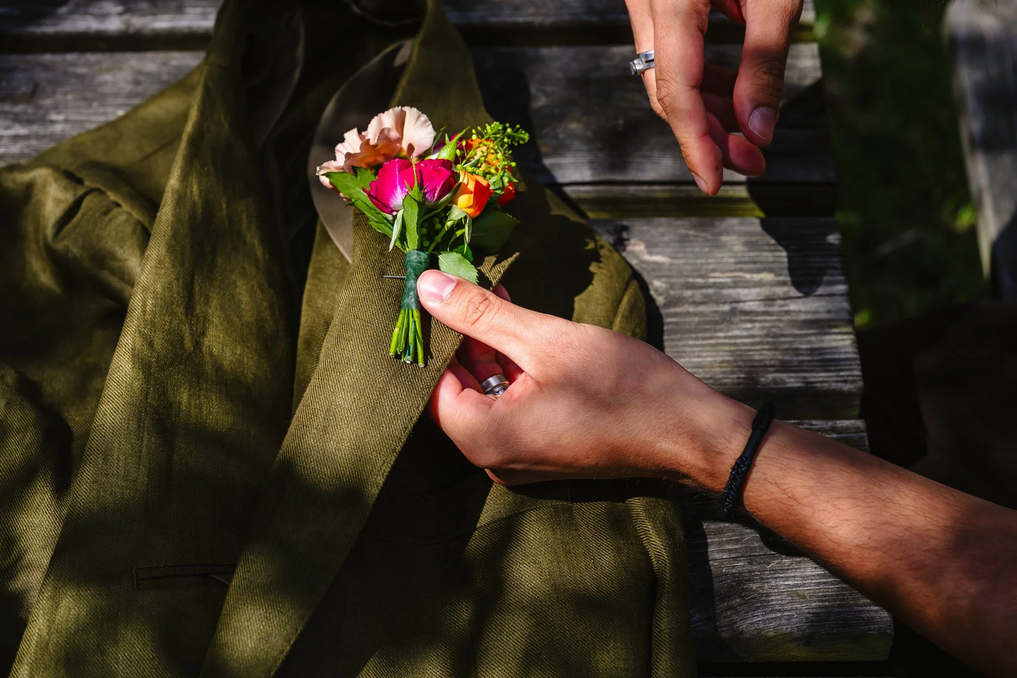  Close up photo of hands putting a floral buttonhole on a green jacket in dappled shade 