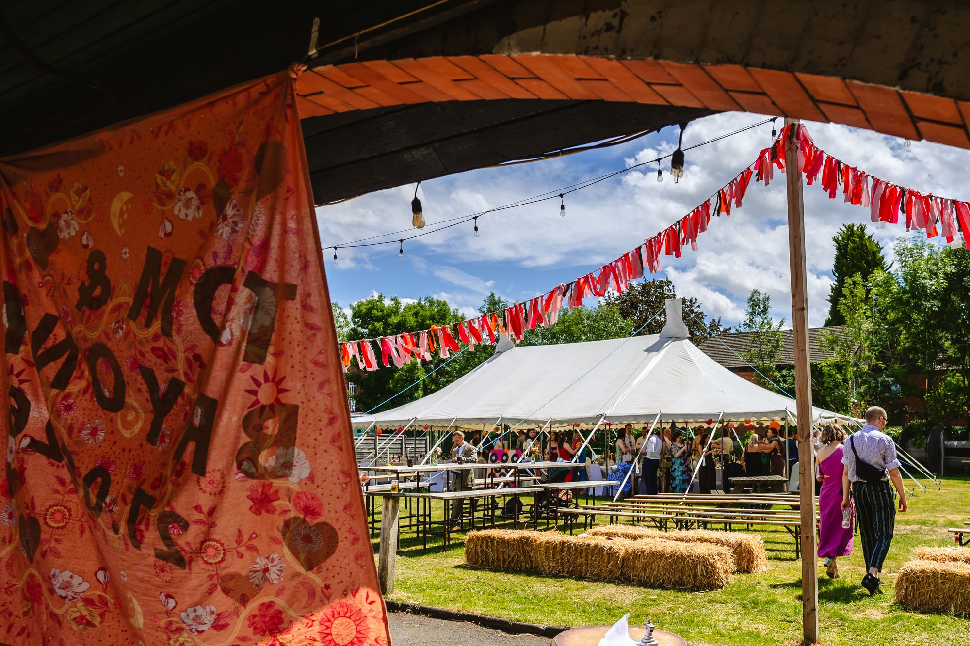  A photograph of the outdoor ceremony space at the Klondyke showing the hay bales, some colourful bunting and the marquee in the background. There’s also a decorative hanging in the foreground. 
