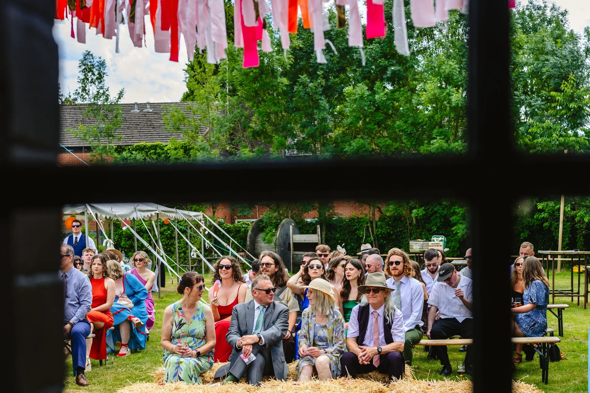  Photograph of the guests at a wedding at the Klondyke, sitting outside on haybales. Photo is taken through a window, with red and pink bunting visible in the top of the frame. 