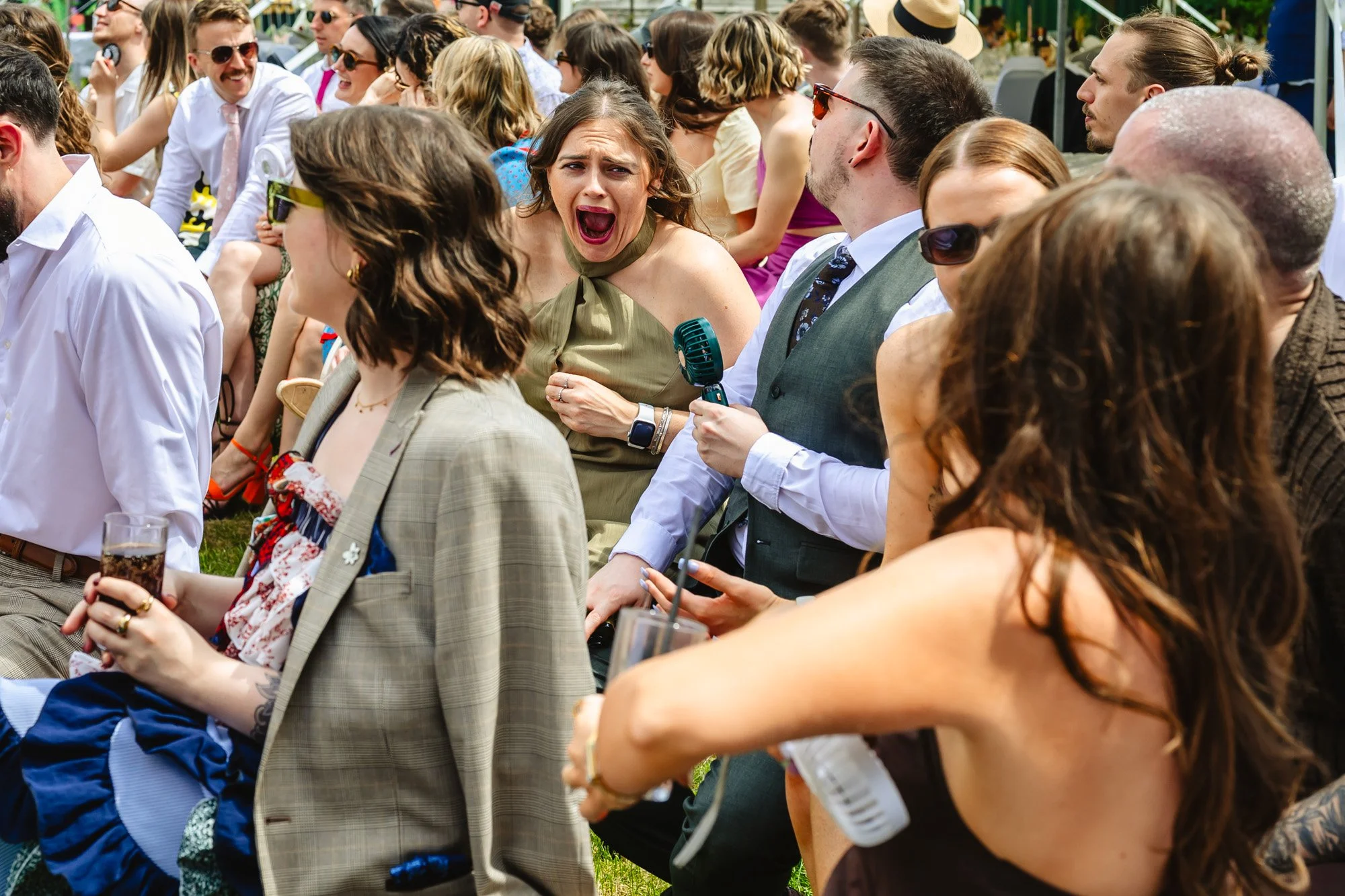  Photograph of a couple of women sat outside at a wedding in a heatwave. One is holding a fan up to her armpit and the other is laughing with her mouth open. 