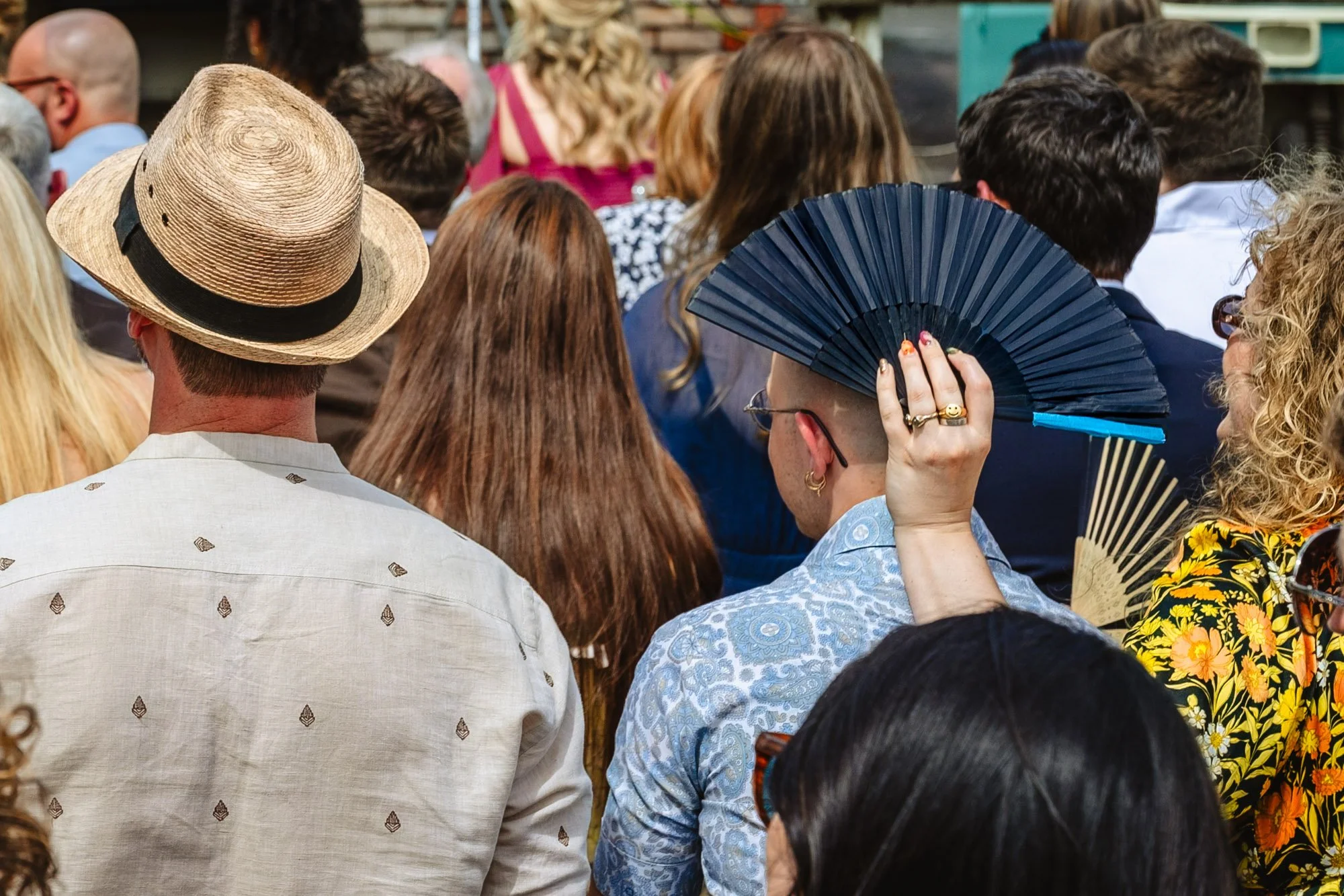  Photograph of a woman holding a black hand fan over a man’s head during a sunny outdoor wedding ceremony 