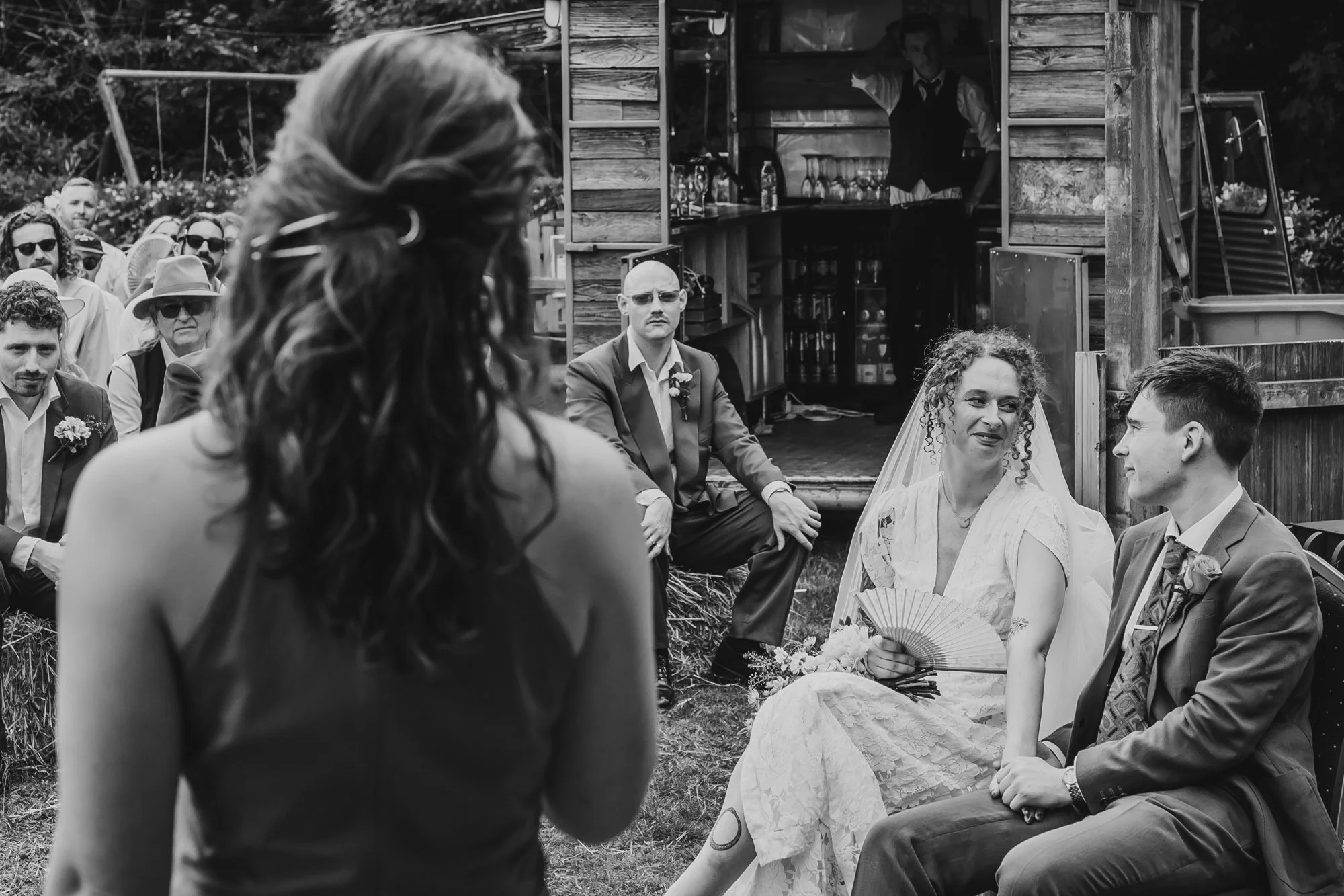  black and white image of a couple looking lovingly at one another while one of their guests performs a reading at their wedding ceremony 