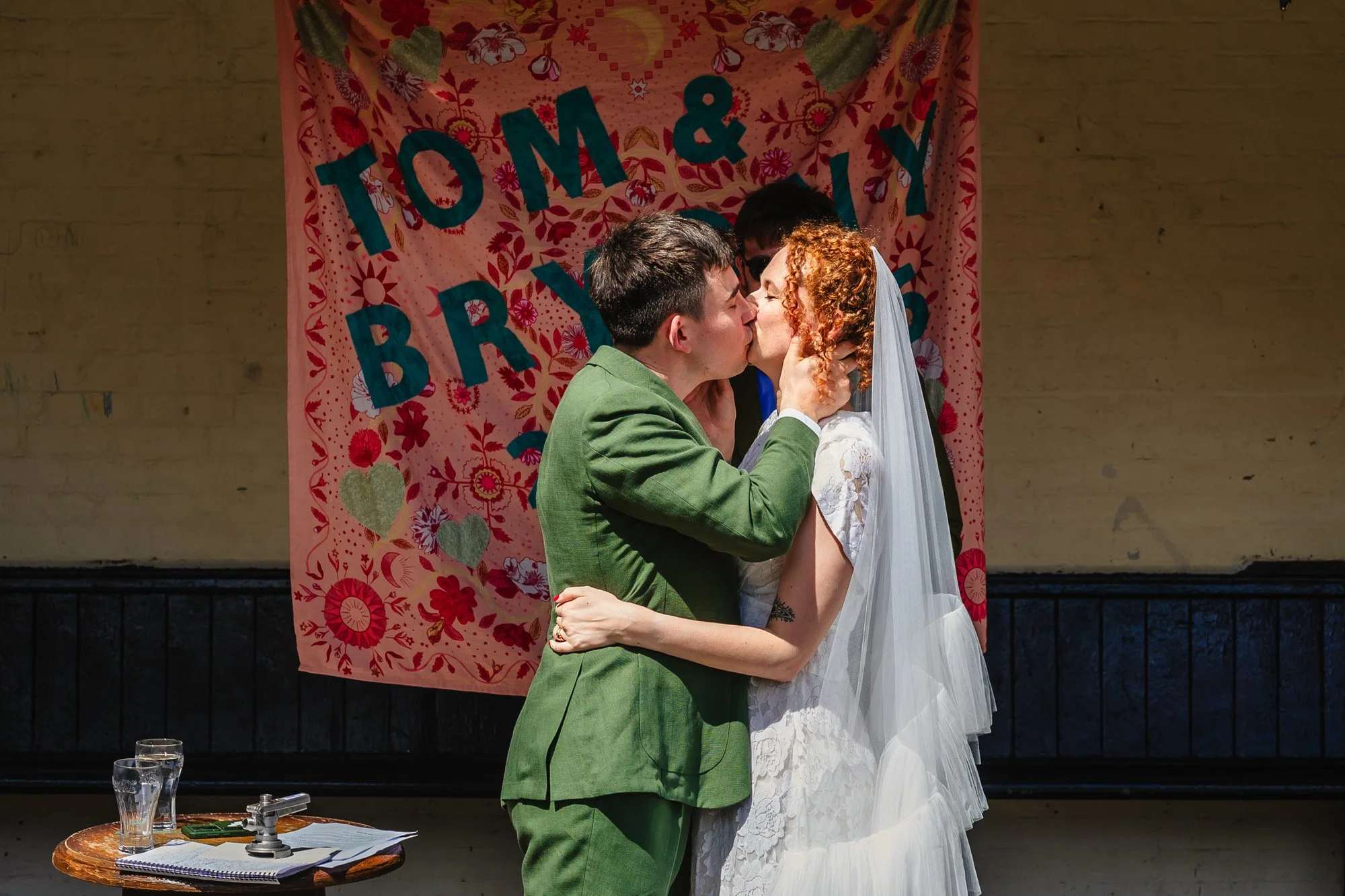  A couple have their first kiss during their outdoor wedding ceremony in Manchester, with their custom pink wall hanging behind them 