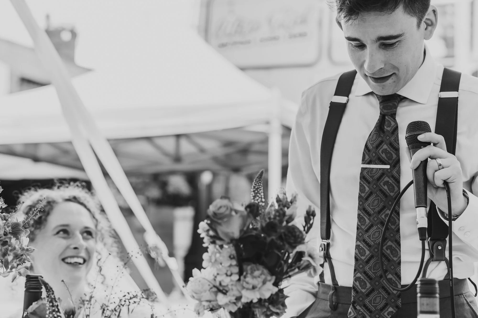  Close up black and white photograph of a man in a tie and braces giving a speech at his wedding breakfast in Manchester. His wife looks at him, smiling. 