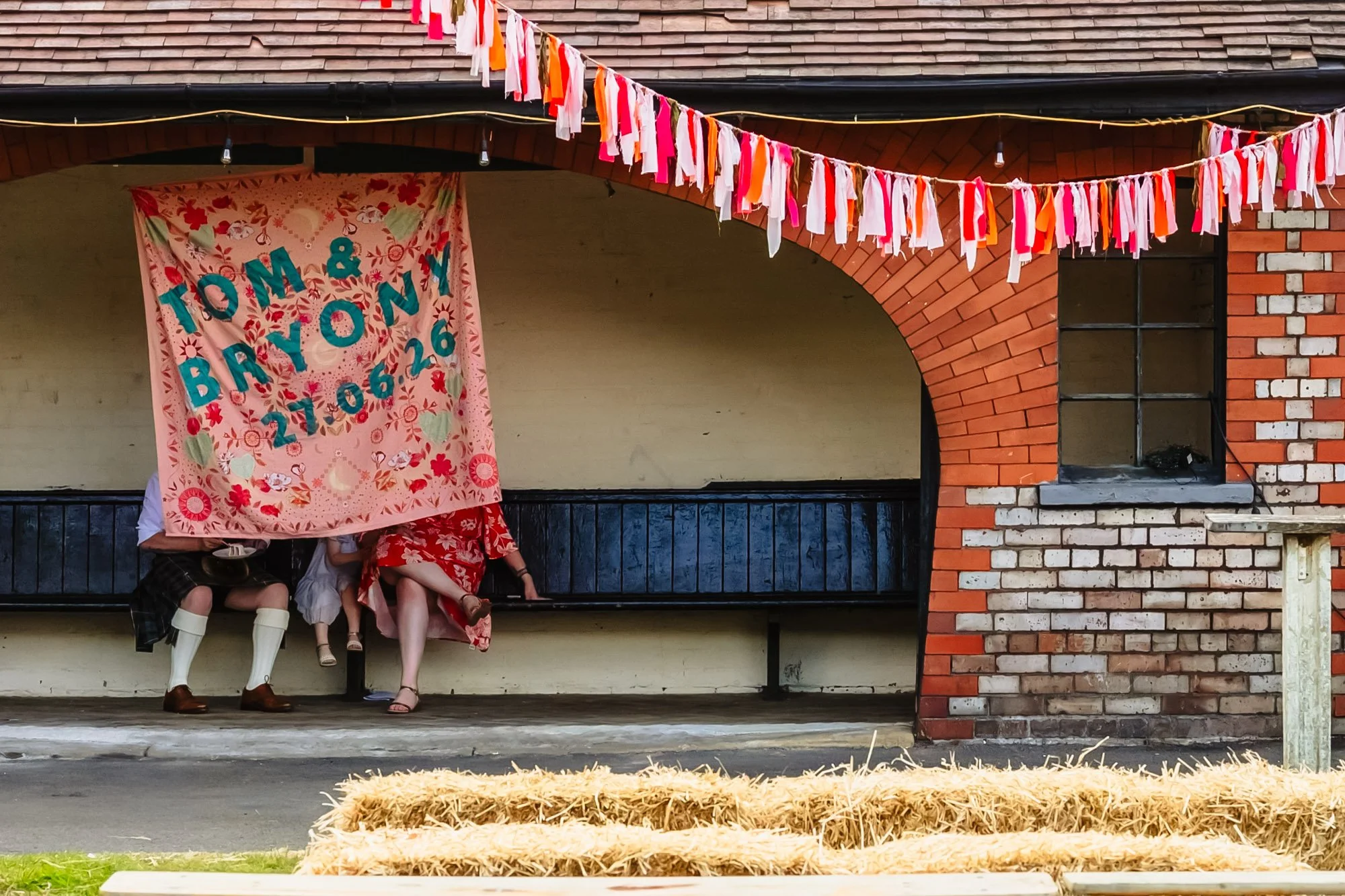  Three people are sitting on a bench behind a hanging embroidery that reads “Tom &amp; Bryony 21-06-26”. Their legs are the only visible part of them. 
