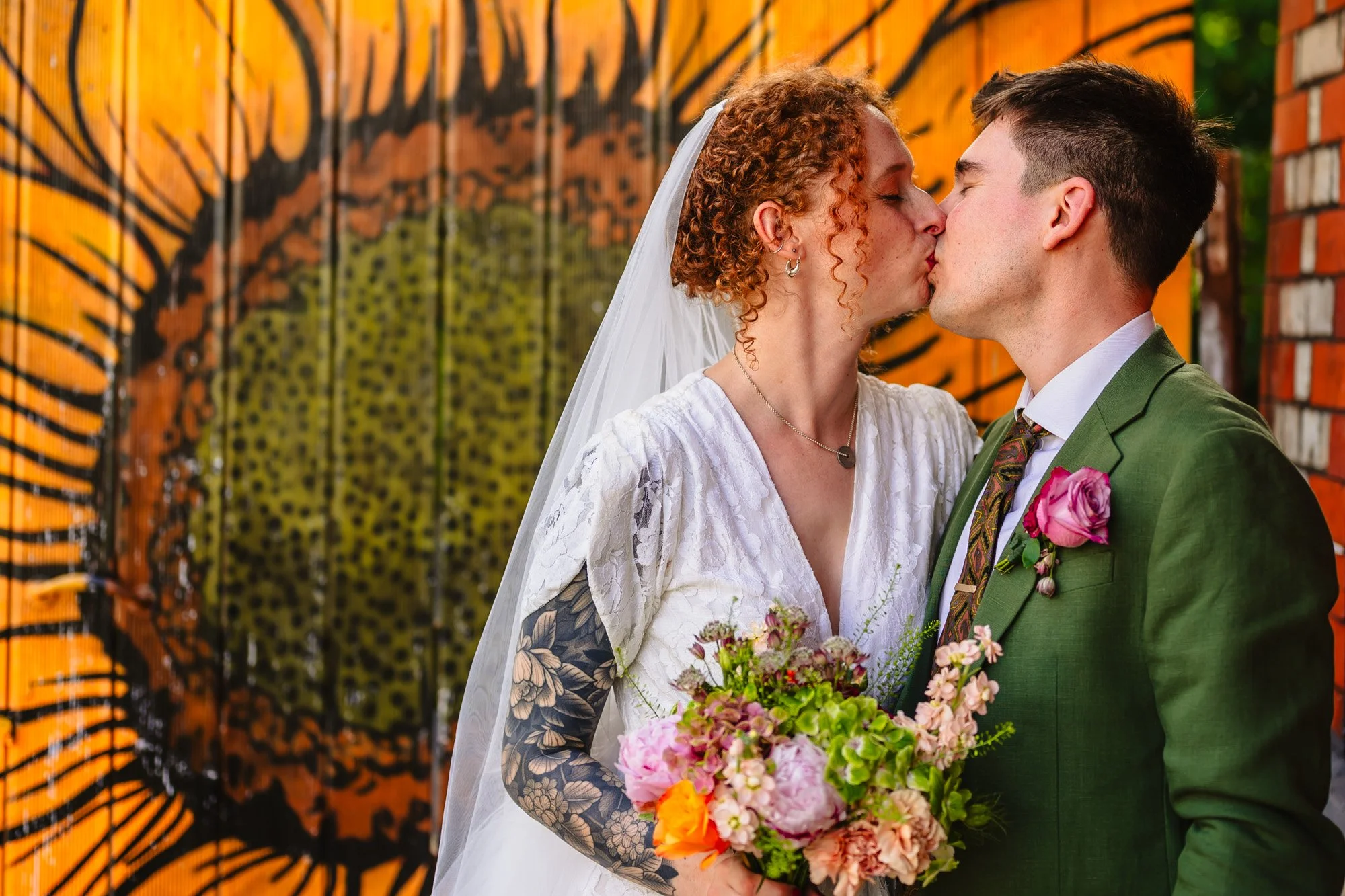  An alternatively styled bride and groom kiss in front of wooden door painted with a huge sunflower. She has curly red hair and a full sleeve tattoo, and is wearing a white lace dress with a veil. He is in a green suit with a pink flower buttonhole. 