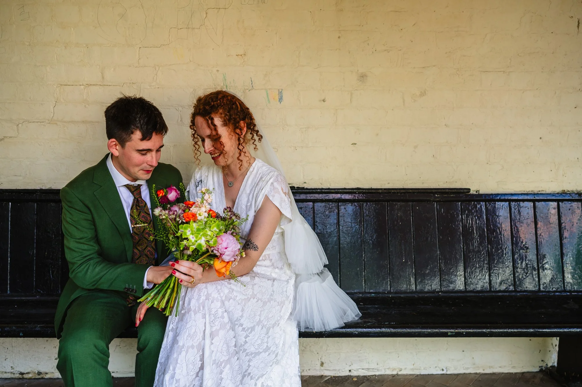 A newly married couple are sitting on a black bench with a whitewashed wall behind them in their wedding clothes 