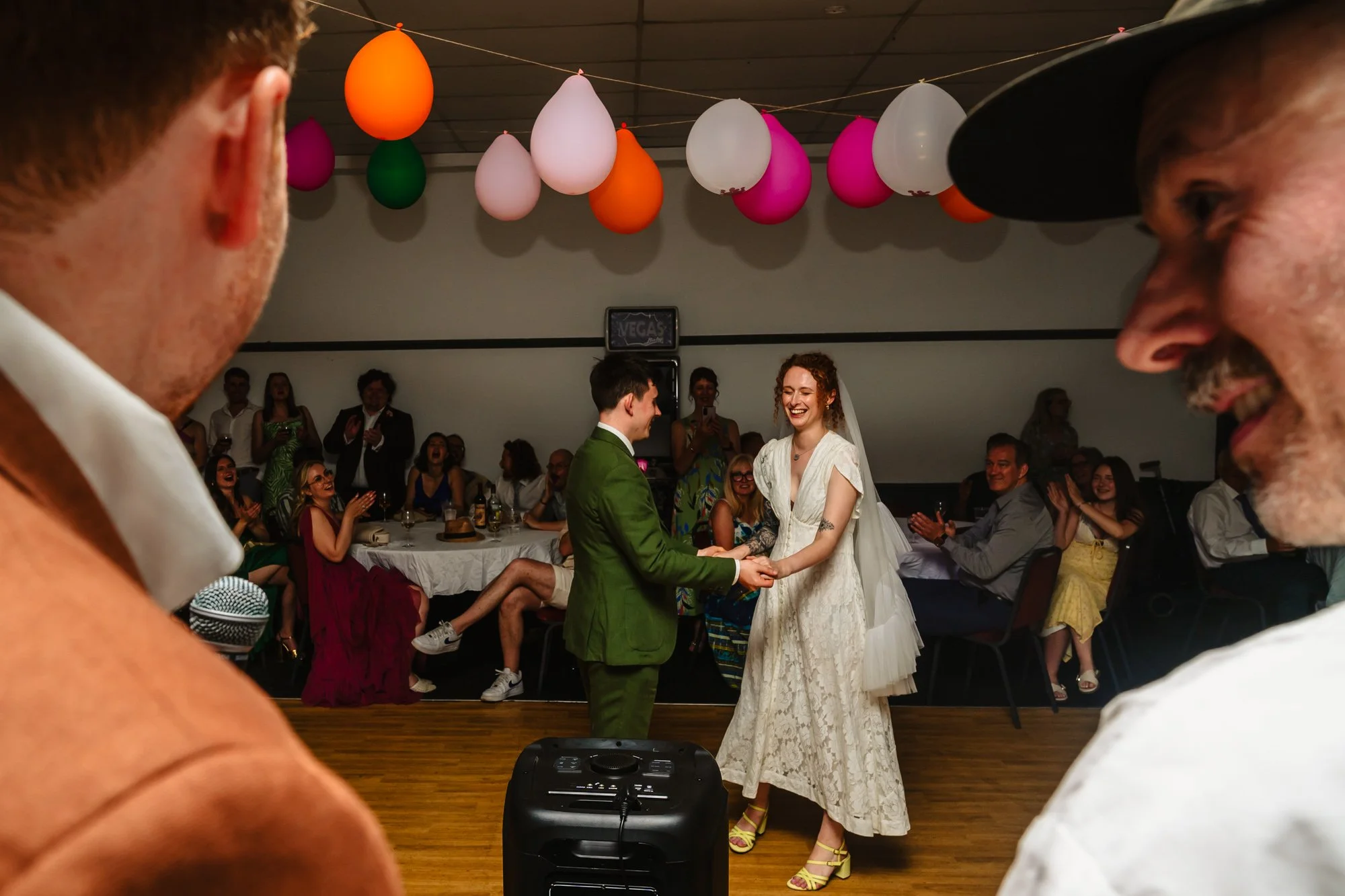  A man and woman begin their first dance at the Klondyke, Manchester. They are framed by the faces of two men in the foreground, with a string of colourful balloons hanging above them. 