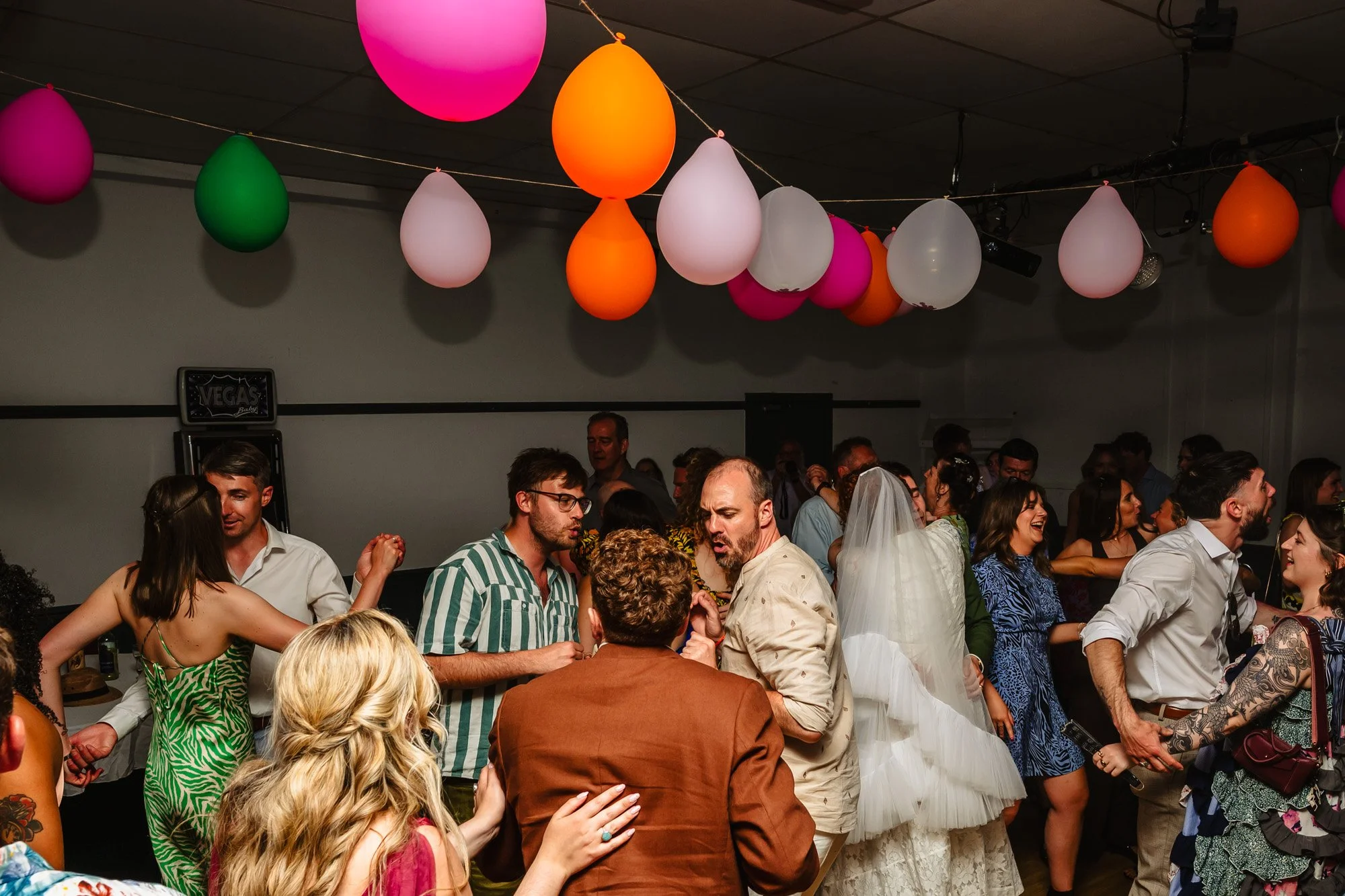  Photograph of guests dancing at a wedding in a spartan room with a string of balloons on the ceiling. 
