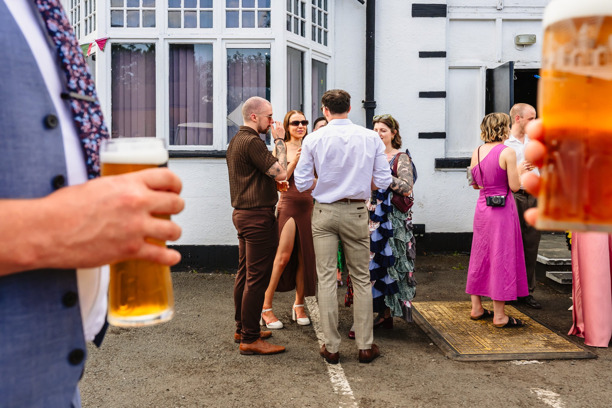  photograph of wedding guests standing outside drinking at a reception, framed by two men holding pints of beer either side of the frame 