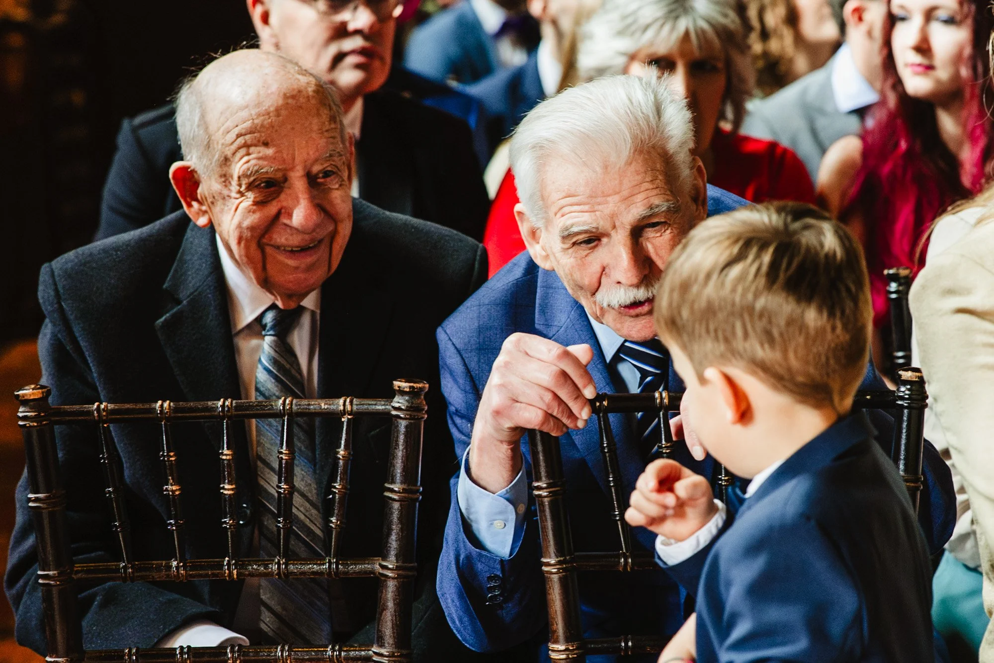 Documentary wedding photography moment of two older men smiling and playing with a young boy sat in front of them moments before the ceremony is due to start