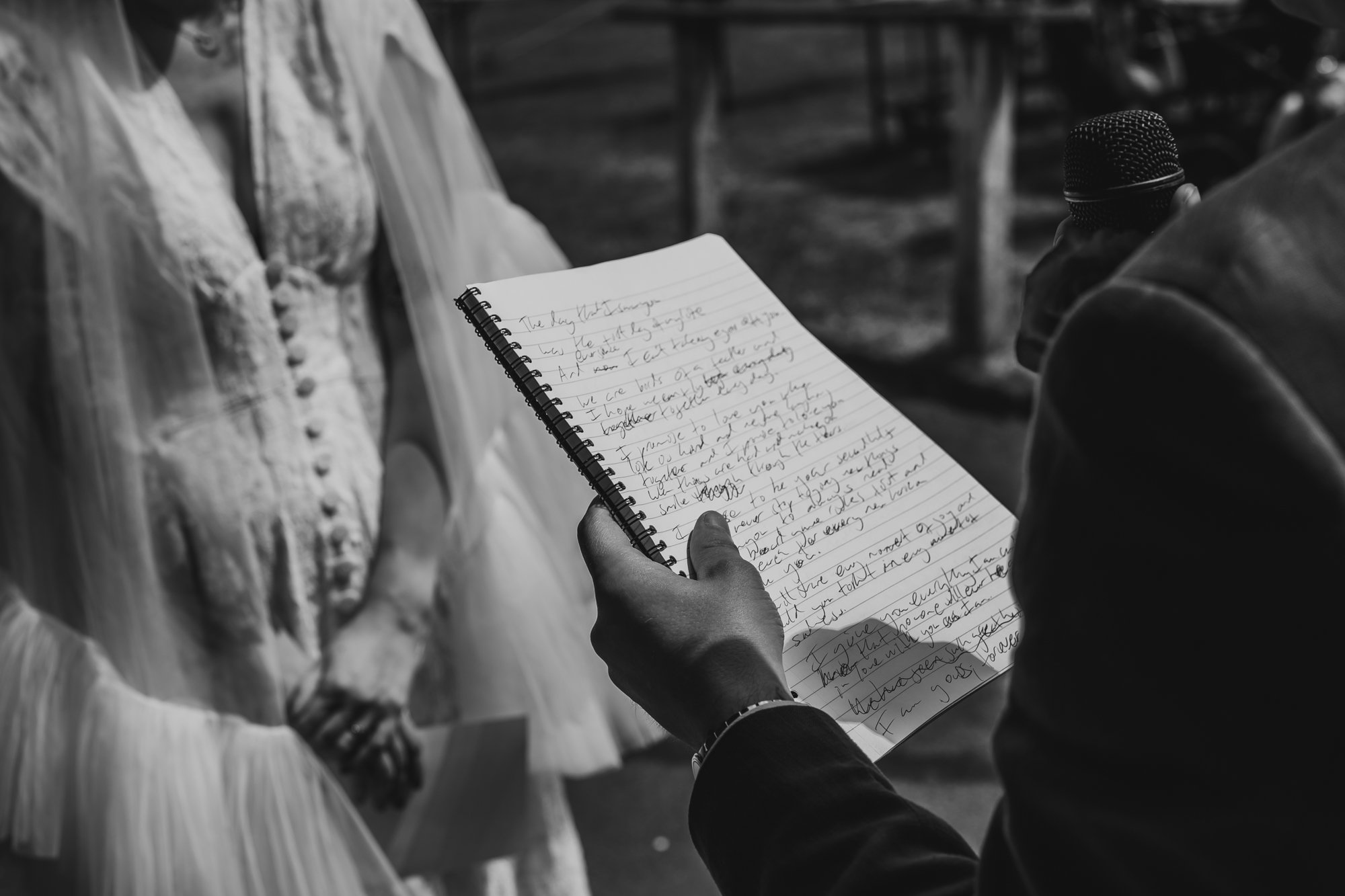Black and white photo of a man holding writing pad. His marriage vows are messily written. In the background a woman in a white dress and veil is visible.
