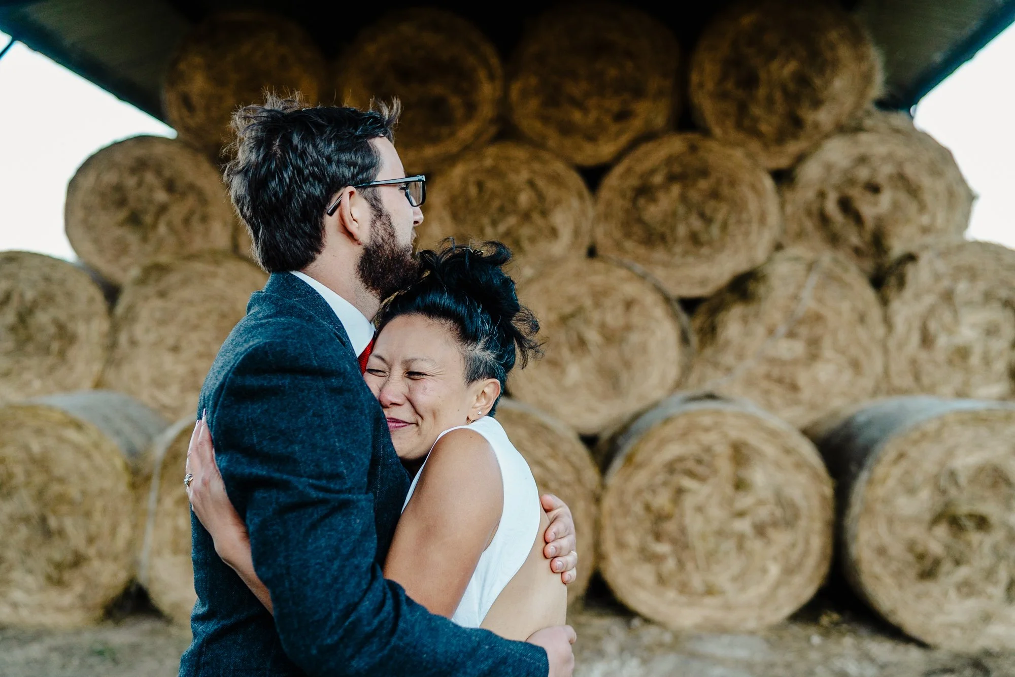 Photograph of a bride and groom having a cuddle outside in front of bales of hay. The bride looks extremely happy. Photo taken at a farm wedding venue in Cheshire