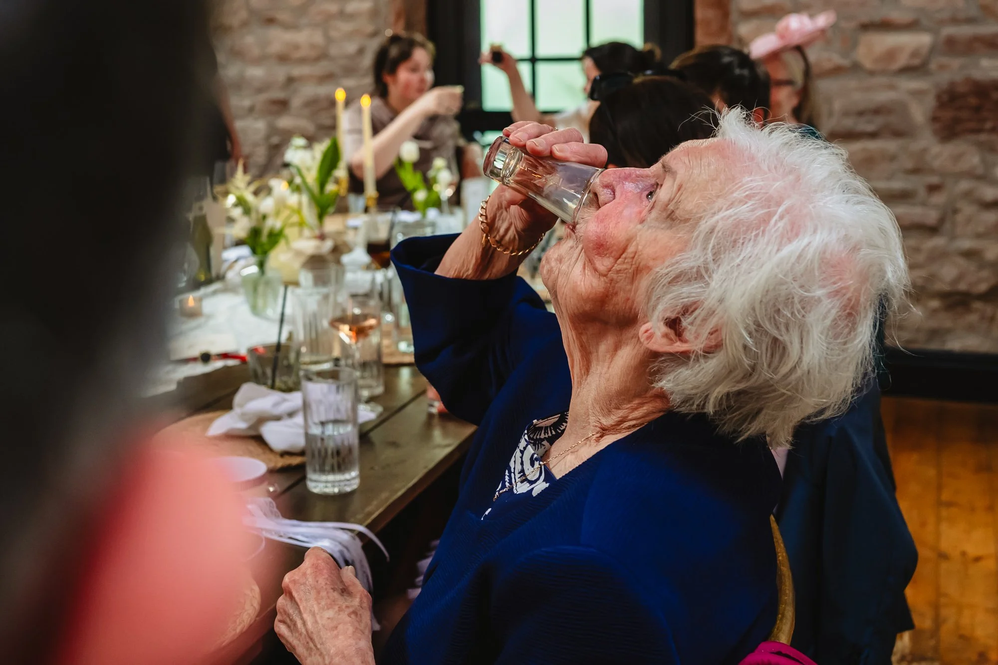Photograph of an elderly white haired woman downing a shot of alcohol while people in the background can be seen toasting. Photo taken during speeches at a wedding at Lost wedding venue in Wirral
