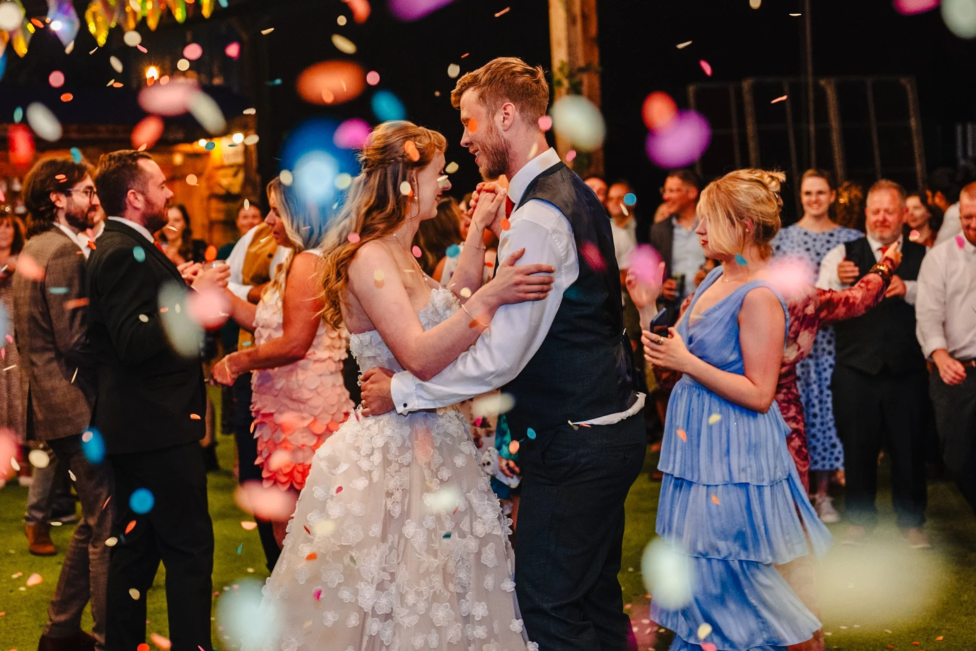 Unposed photograph of bride and groom dancing outside. Their guests are in the background and confetti falls over them. Photo taken at Whitebottom Farm in Stockport, Cheshire.