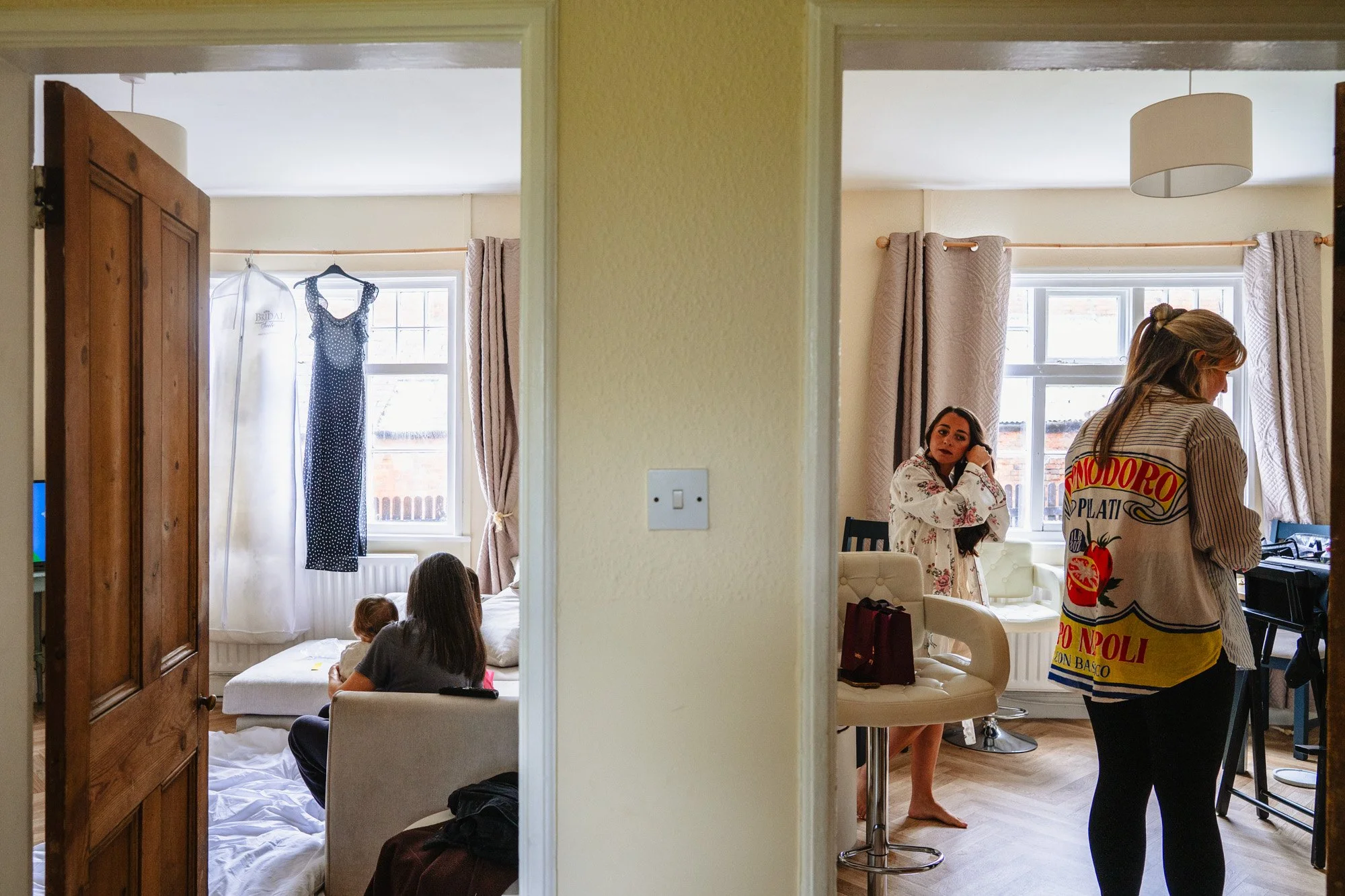 A photograph showing two rooms. In one, a woman sits with two young children, with a wedding dress and a blue spotted dress hanging by the window. In the other a woman in a floral dressing gown puts her earrings in.