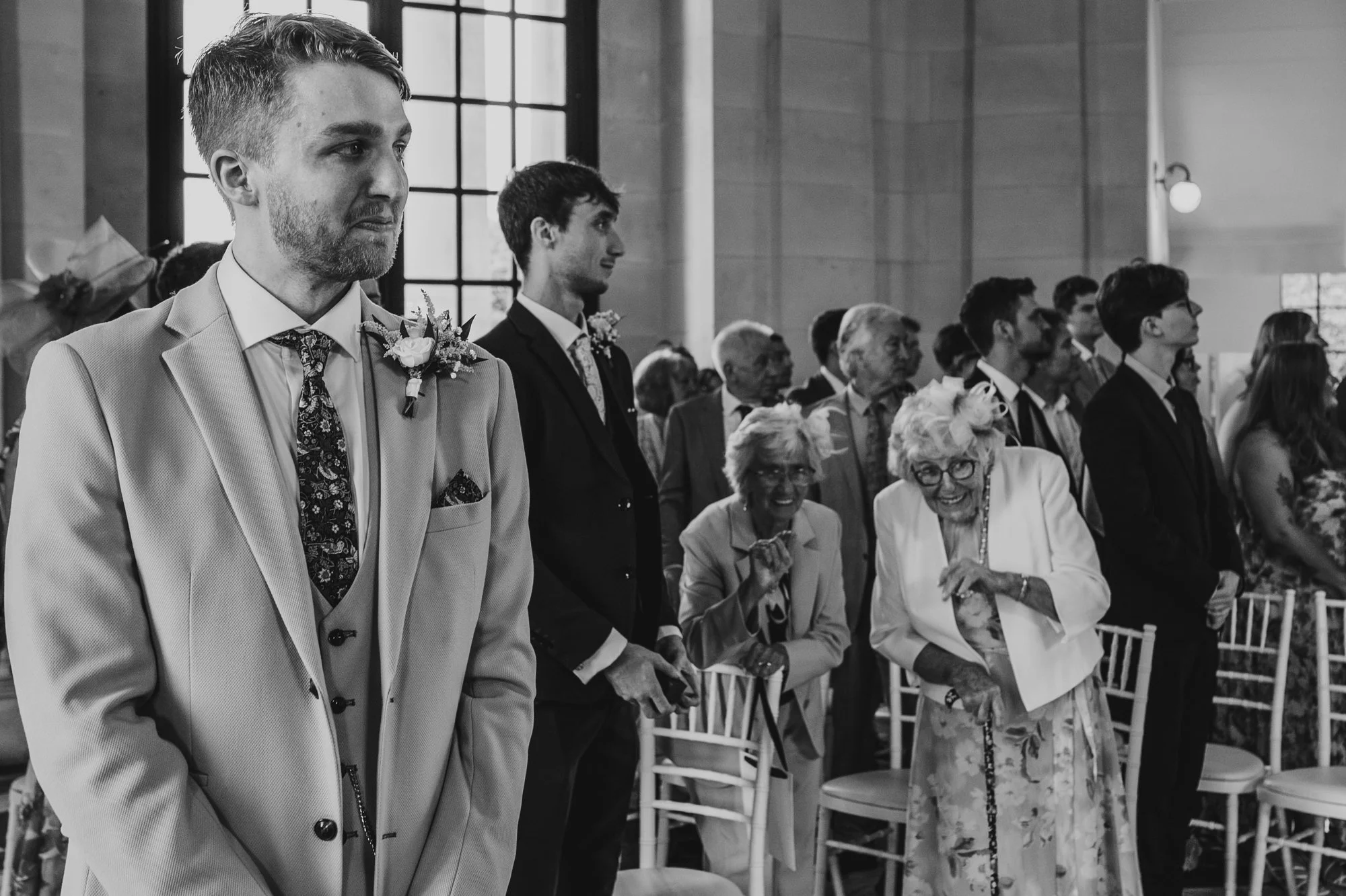 photograph of a groom looking emotional during a wedding ceremony while guests stand in the background. Two elderly ladies are smiling and looking in a different direction to everyone else.