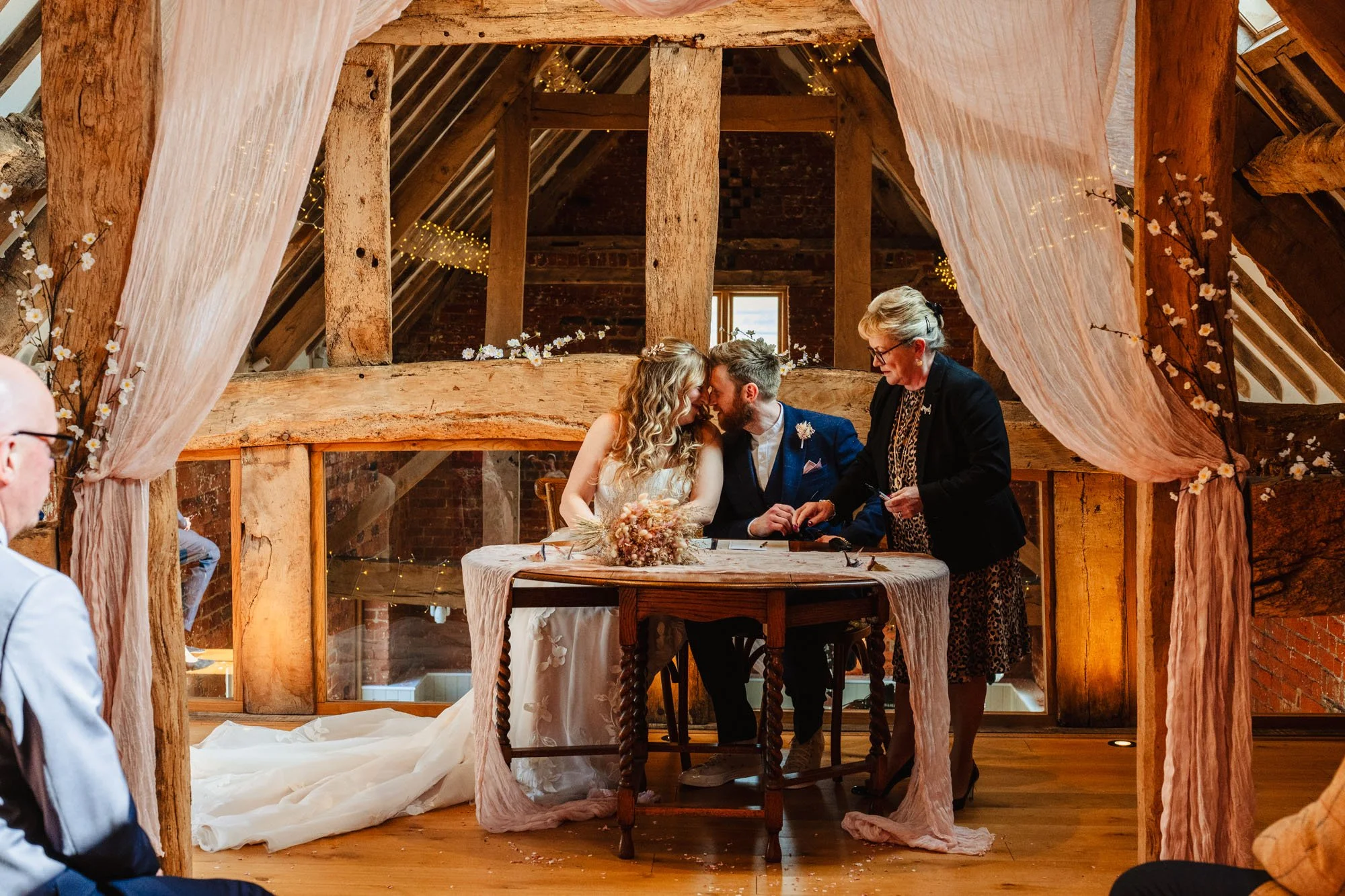 photograph of a couple sitting down and nuzzling their faces together during their wedding ceremony at Moat Hall barns in Shropshire. They are oblivious to the registrar trying to move the paperwork near them.