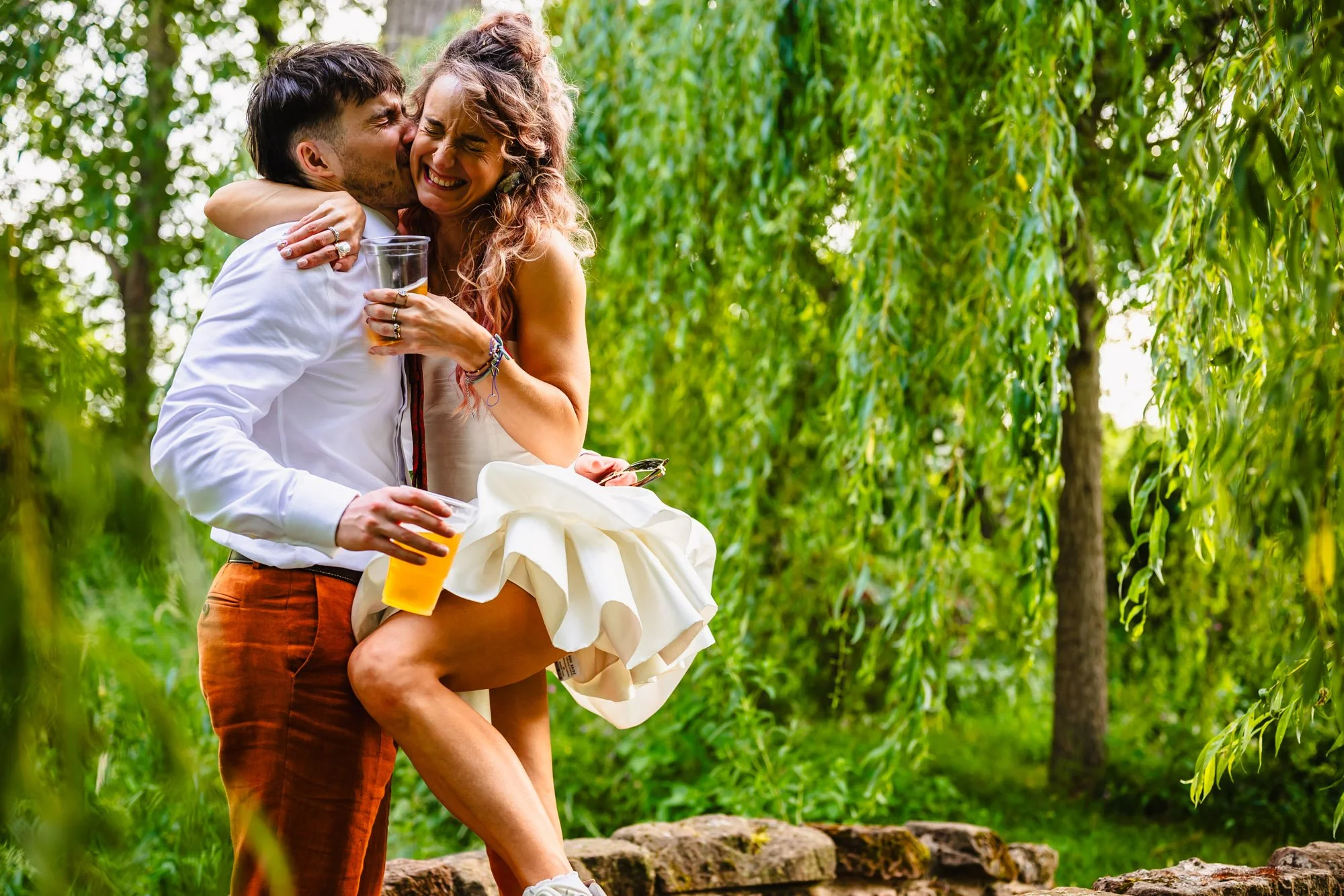 A bride and groom have a hug on a bridge at Duddon Mill Farm with a weeping willow in the background. They're both holding a cup of beer and squeezing their eyes closed while they smile. He is kissing her cheek.