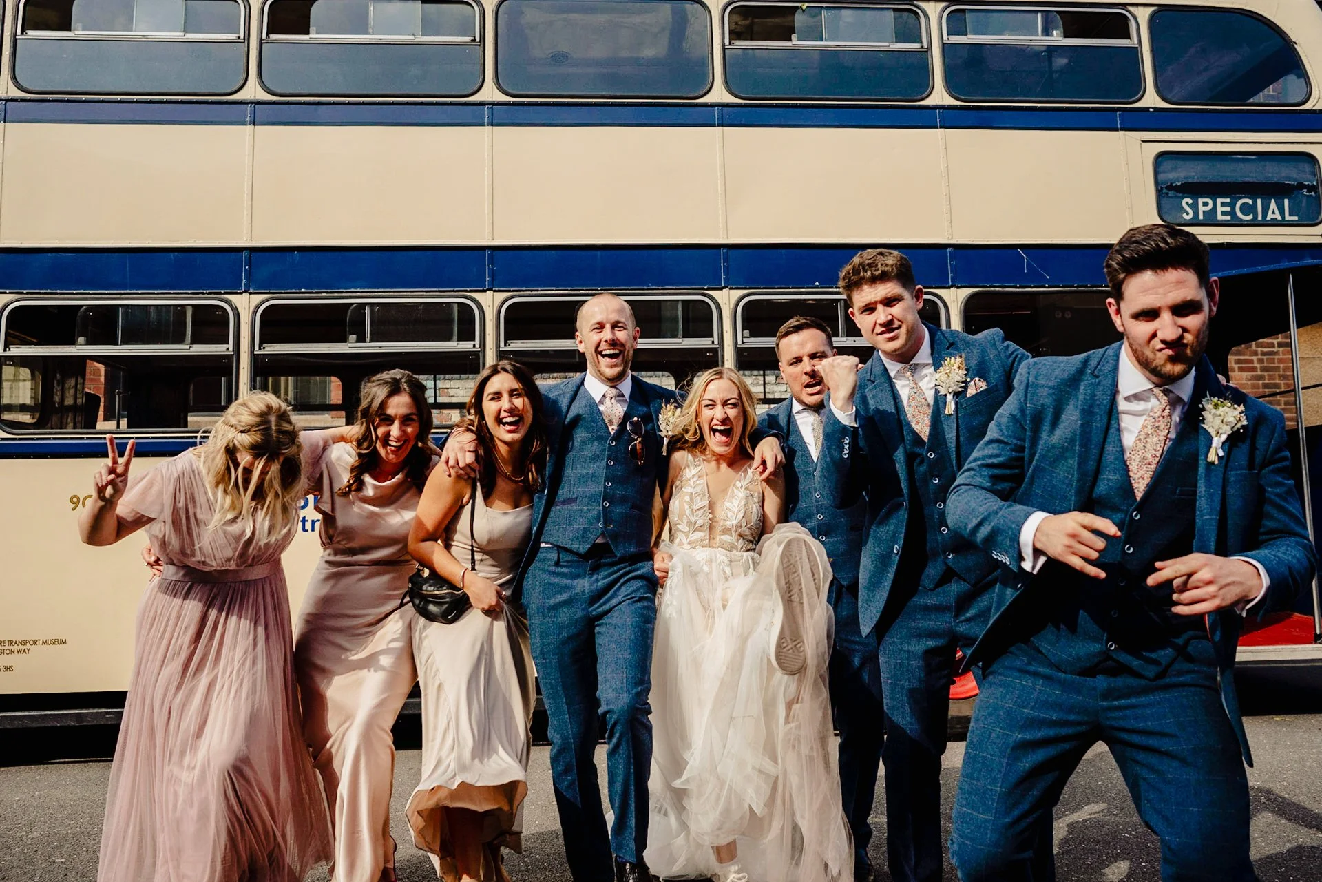 Group photo of bridesmaids and groomsmen all with their arms round each others' shoulders, doing a funny walk and laughing. There's an old routemaster bus behind them. Taken in Kelham Island, Sheffield.
