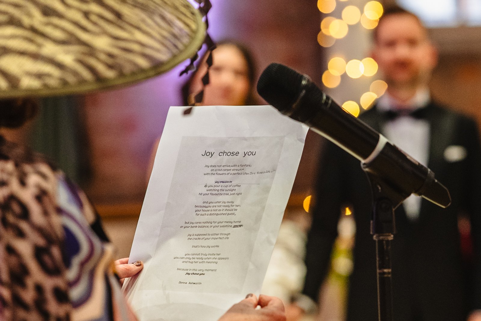 close up photograph of a woman holding a wedding reading called Joy Chose You with a microphone in the foreground and a bride and groom in the background by documentary wedding photographer Caroline Goosey